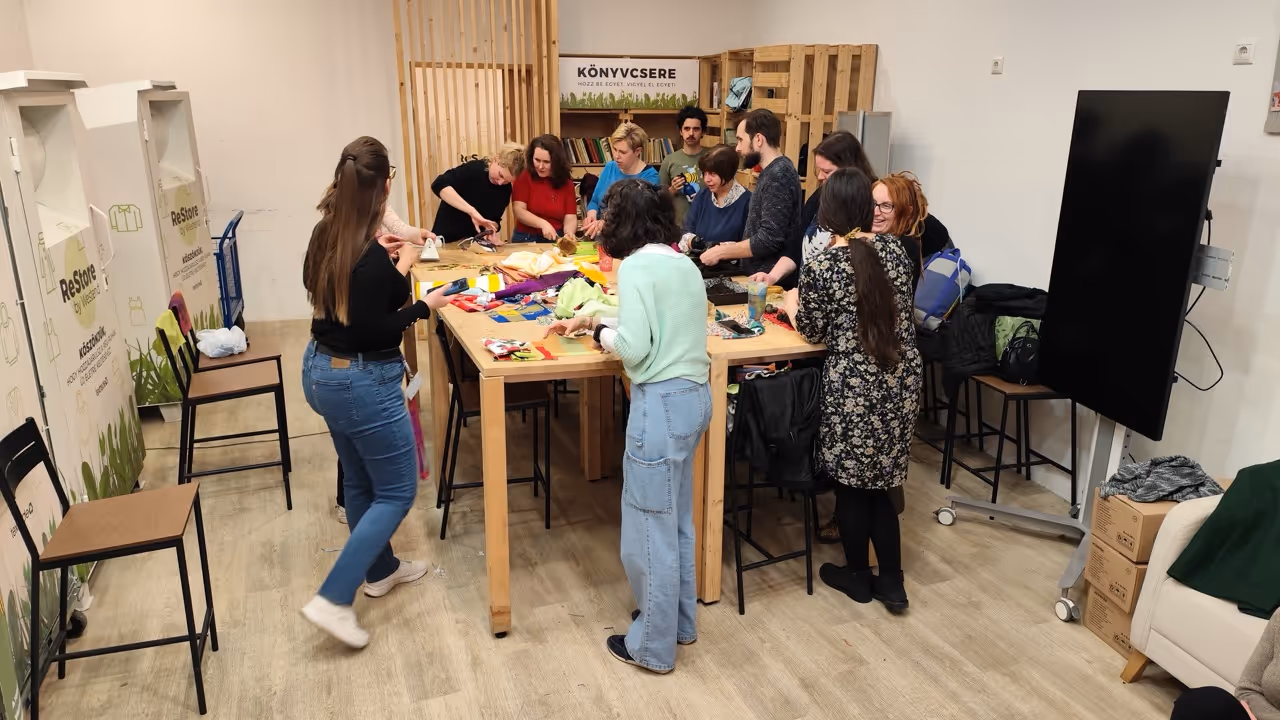 Tizenegy ember egy asztal körül áll egy műhelyben, ahol különféle színes anyagokat és eszközöket használva dolgoznak. A háttérben egy polcon könyvek és egy 'Könyvcsere' tábla látható.