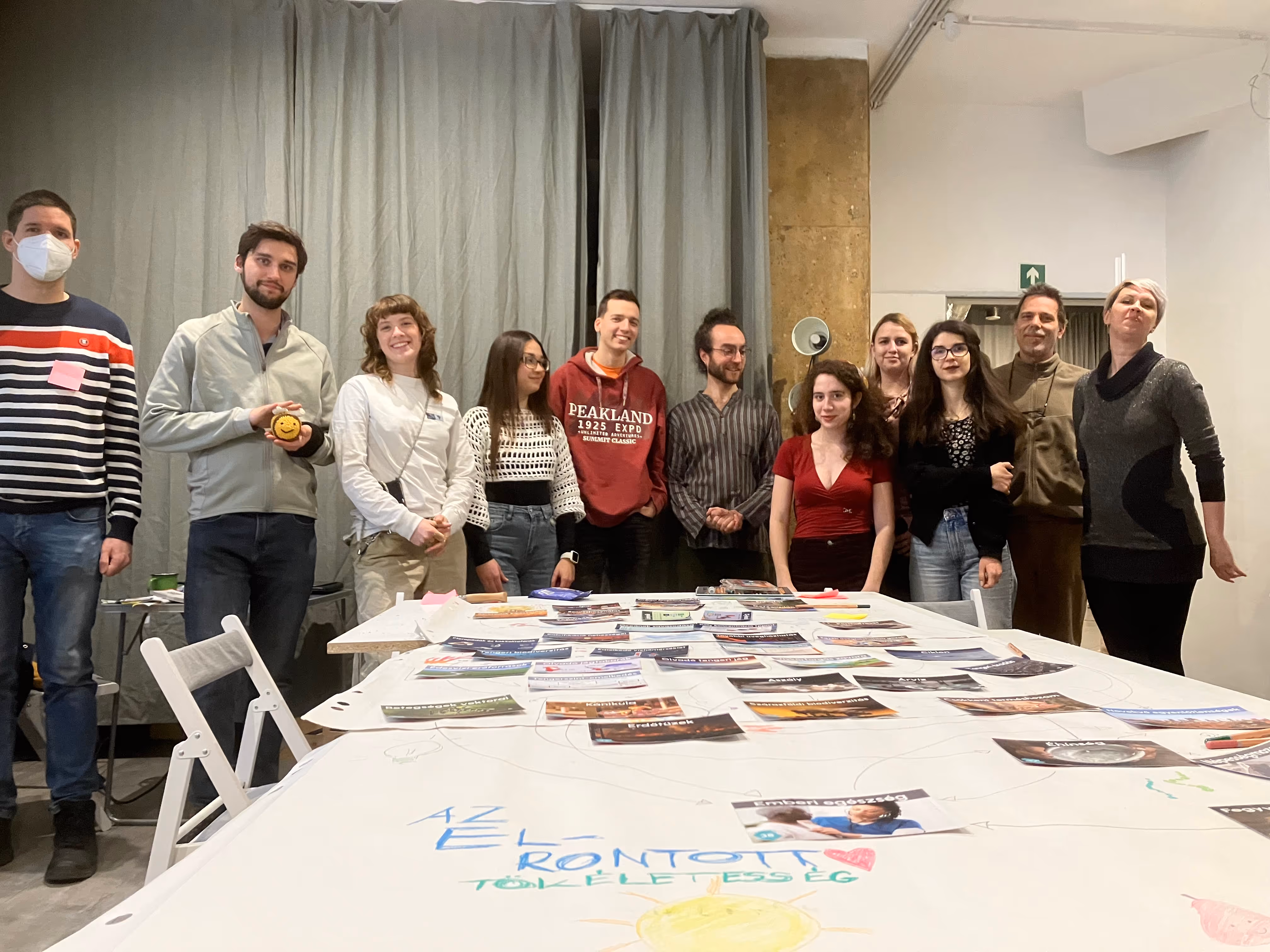 Tizenegy ember áll egy nagy asztal mögött, amelyen papírok és képek vannak szétszórva, a háttérben szürke függöny és világos fal található.