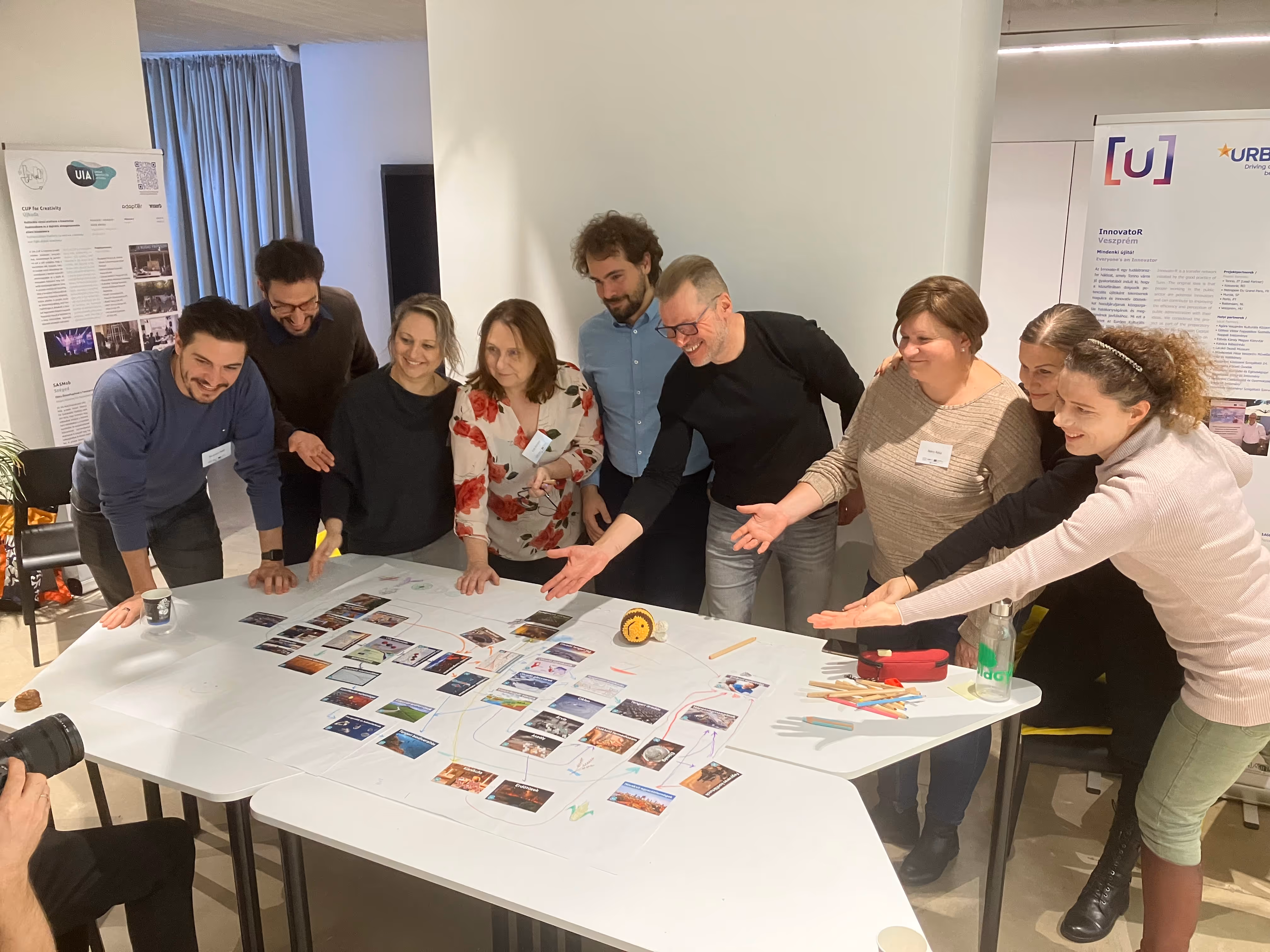 Nyolc ember egy asztal körül áll, amelyen papírra ragasztott képek és rajzok vannak, közösen egy projekten dolgoznak.