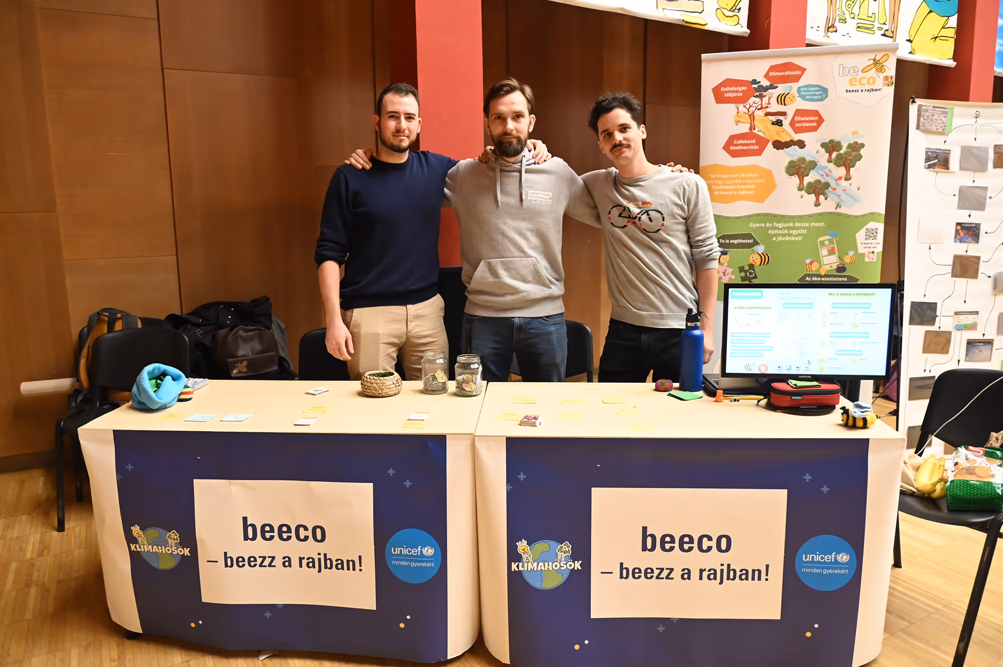 Három férfi áll egy asztal mögött, amelyen poszt-it cetlik és üvegek vannak, az asztal előtt két banner a beeco programról a Klímahősök és UNICEF logóival.