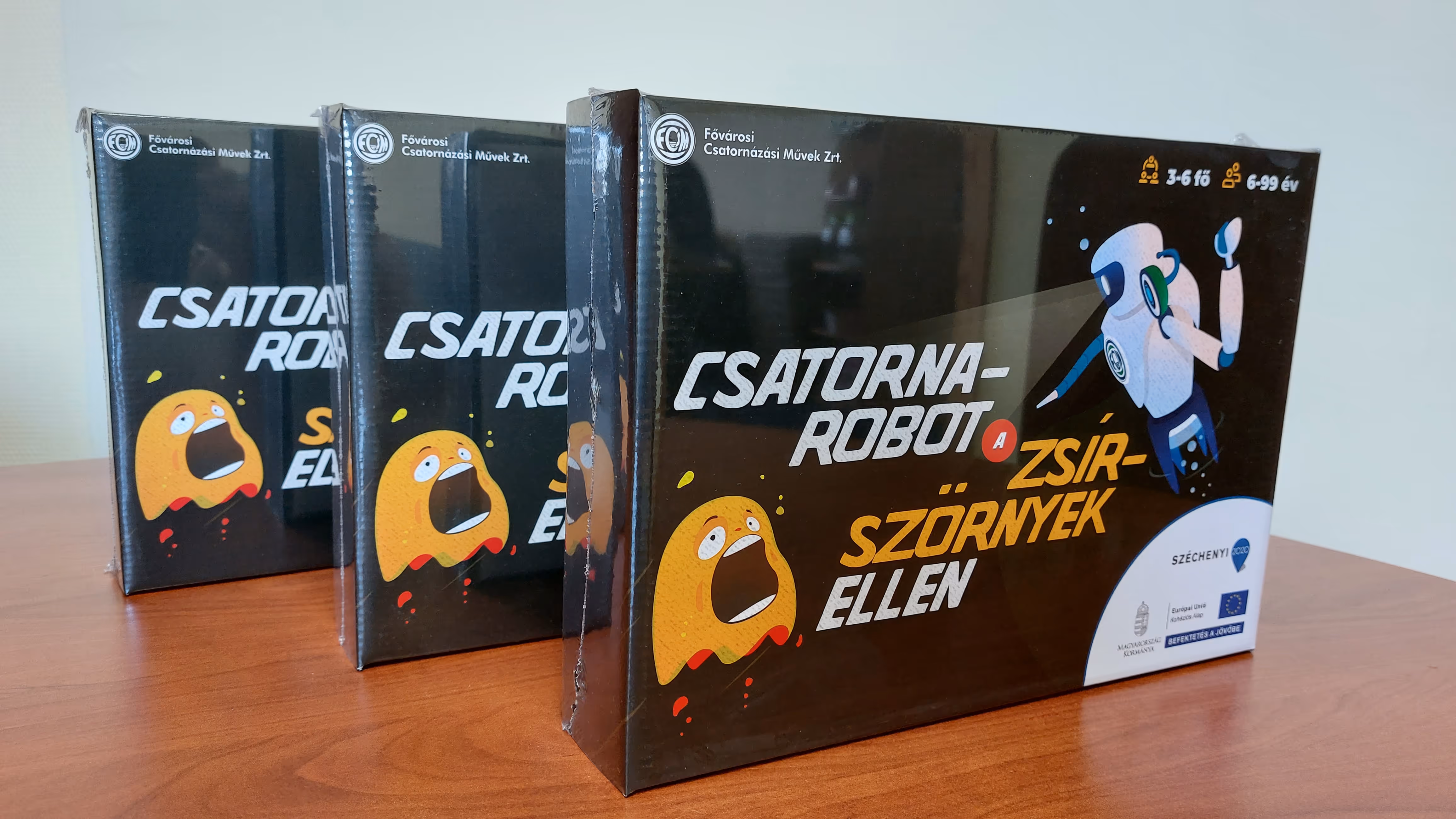 Három Csalorna-robot társasjáték doboz egymás mögött egy fa asztalon, a dobozokon egy robot és egy ijedt sárga szörny ábrája látható.