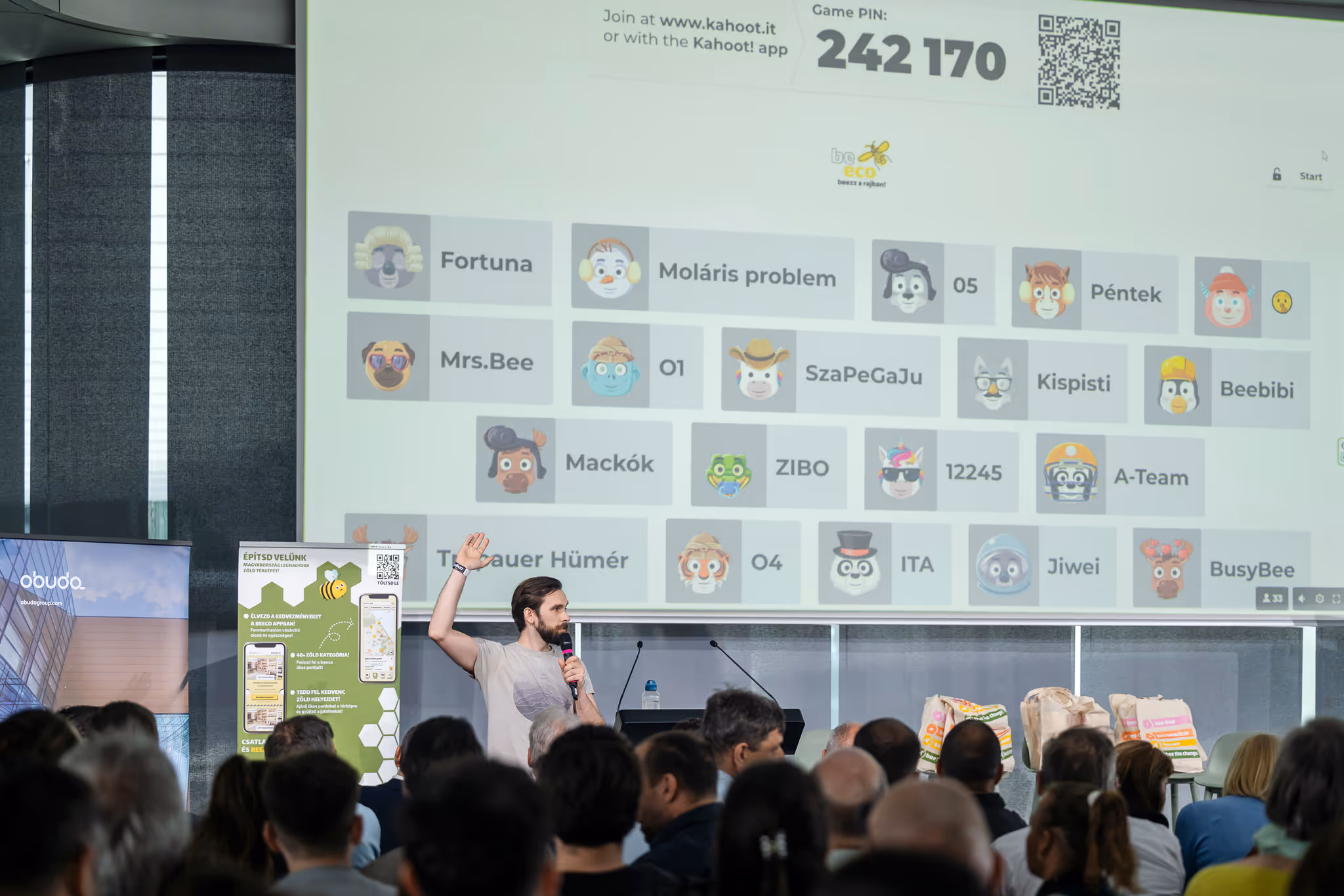 Egy férfi mikrofonnal a kezében előadást tart, míg közönség figyel egy nagy képernyőt, amelyen Kahoot játék résztvevői láthatók.