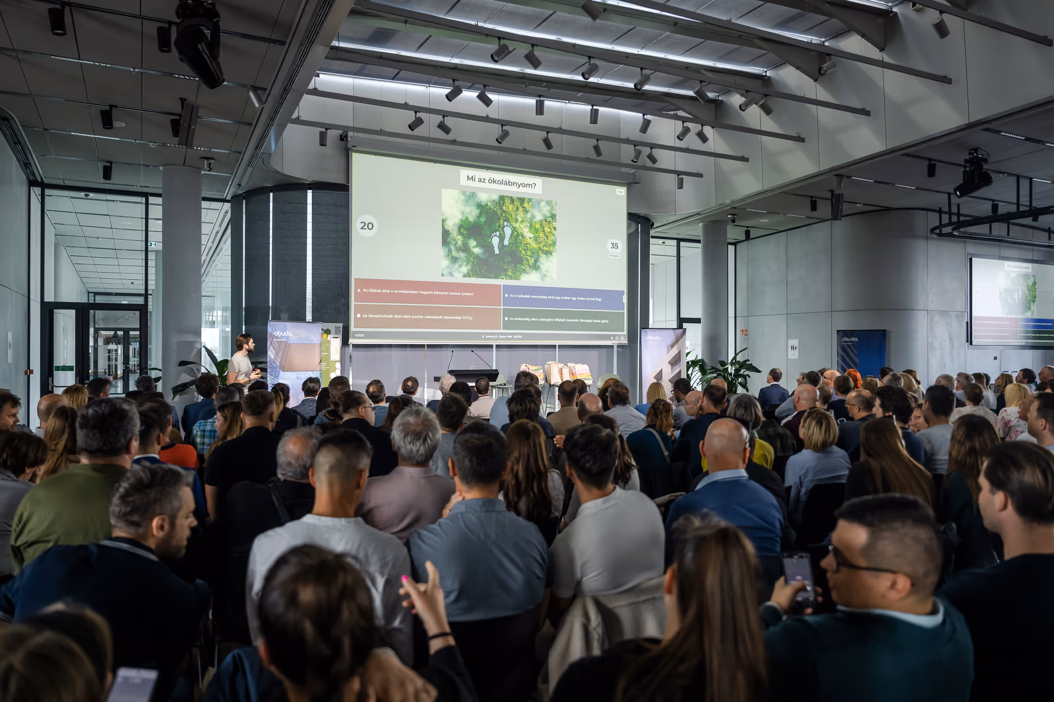 Tömeg figyel egy előadót egy modern konferenciateremben, aki környezeti lábnyomról szóló diát mutat be.