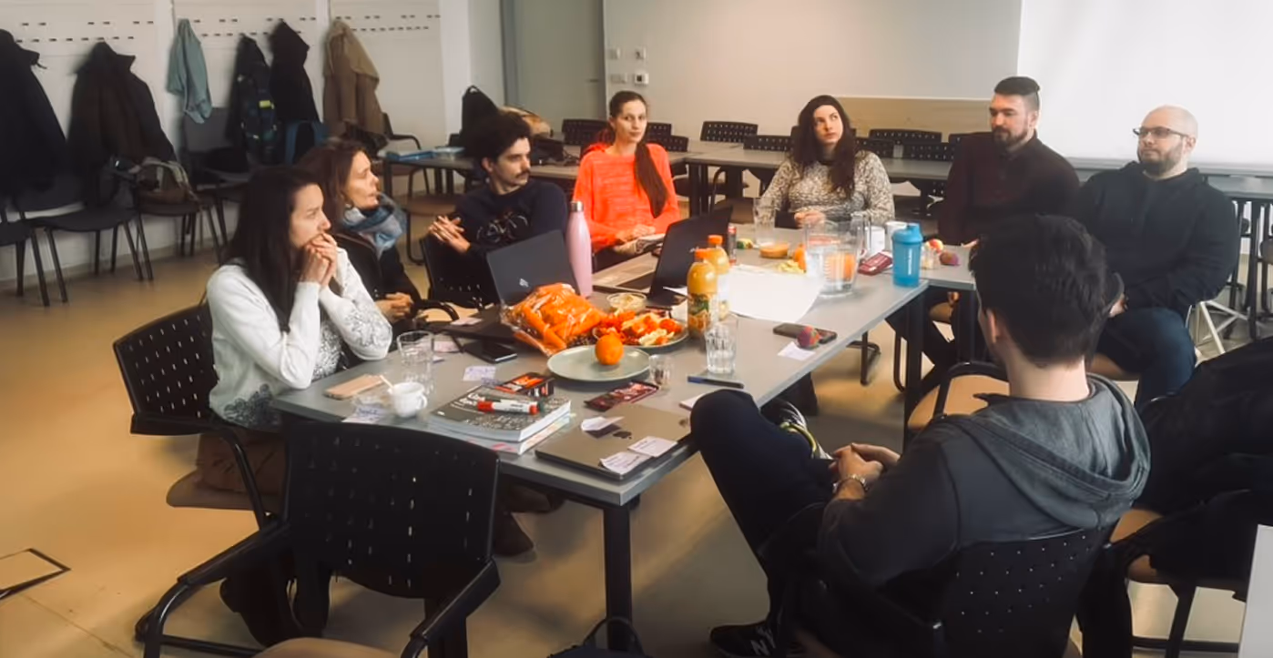 Kilenc ember egy asztal körül ül egy megbeszélésen, asztalon laptopok, italok és harapnivalók.