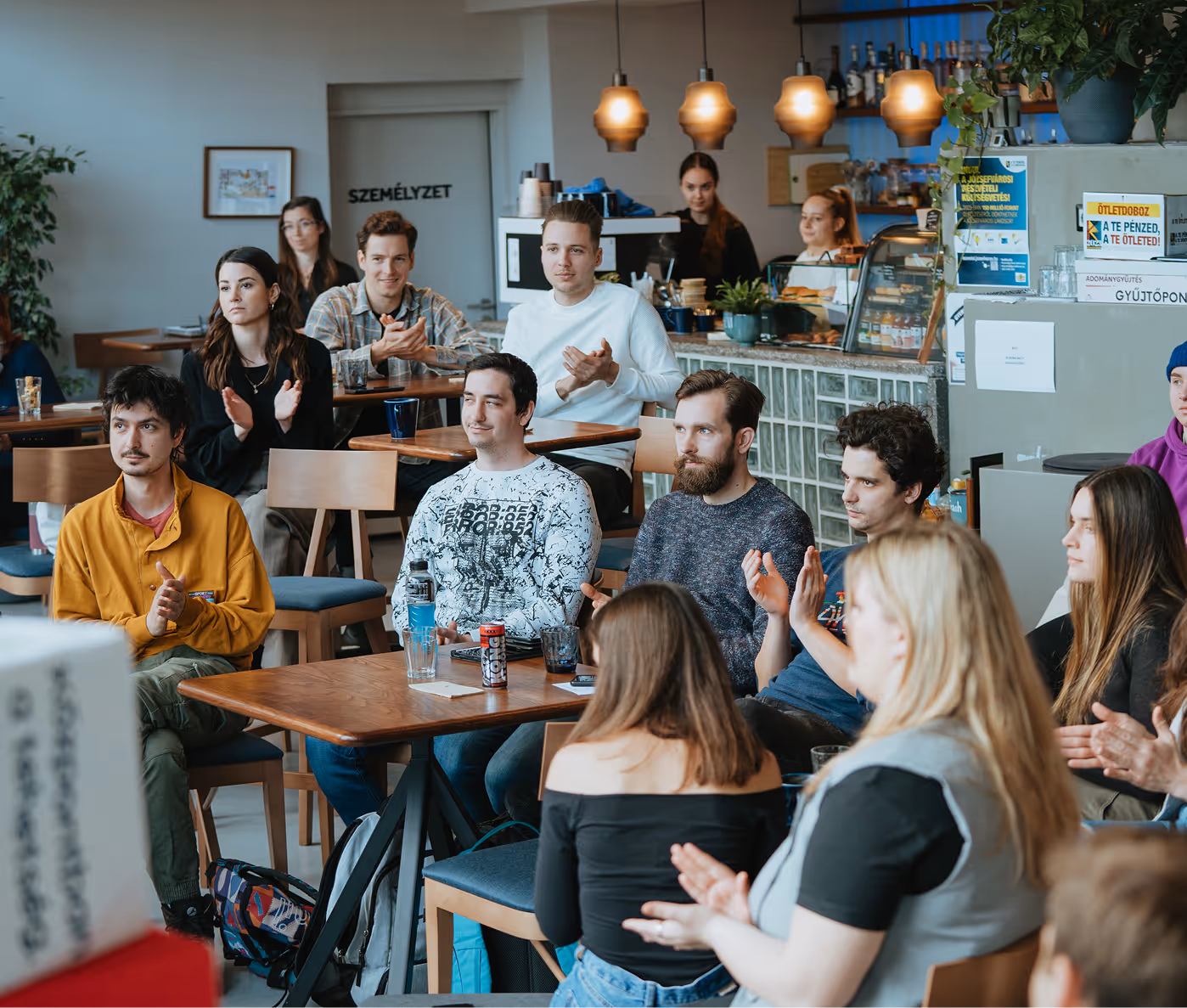 Fiatal felnőttek egy kávézóban ülnek, figyelnek és tapsolnak egy előadáson.