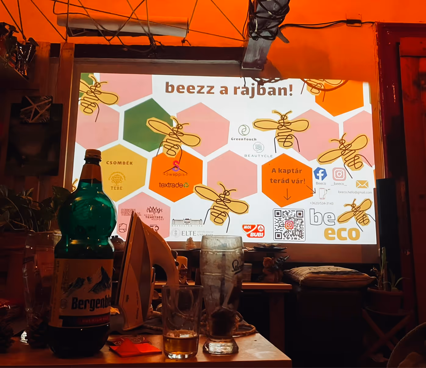 Egy asztalon üres és félig tele sörös poharak, üres sörösüveg és vasaló, háttérben egy világos vásznon méheket és hatszögeket ábrázoló, környezetbarát eseményt hirdető vetítés látható.