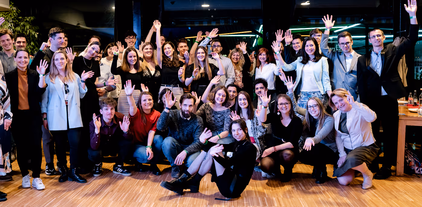 Csoportkép vidám fiatal felnőttekről, akik térdelve és állva, kézfelrázással pózolnak beltéri környezetben.