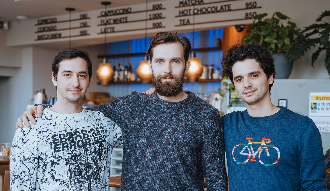 Három fiatal férfi áll egy kávézóban, egyikük szakállas, egyikük fehér pulóvert visel fekete mintával, a harmadik kék pulóvert színes biciklis mintával.