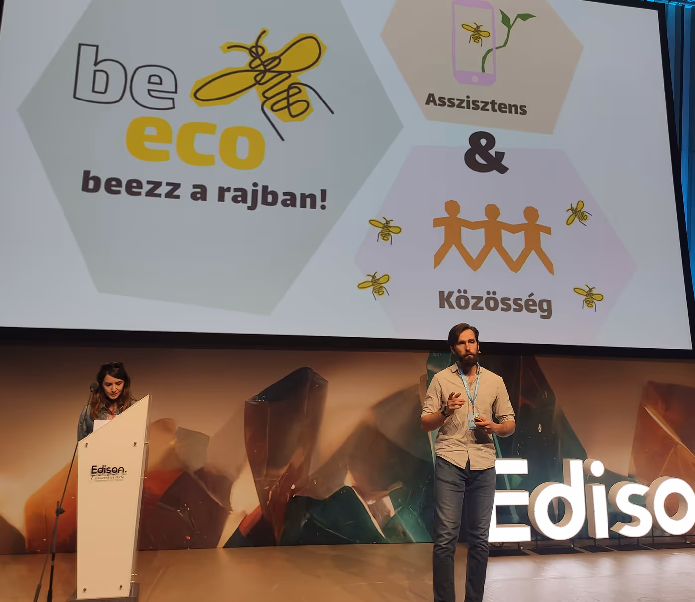 Két előadó egy színpadon egy 'be eco beezz a rajban!' és 'Asszisztens & Közösség' feliratokat tartalmazó prezentáció előtt az Edison konferencián.