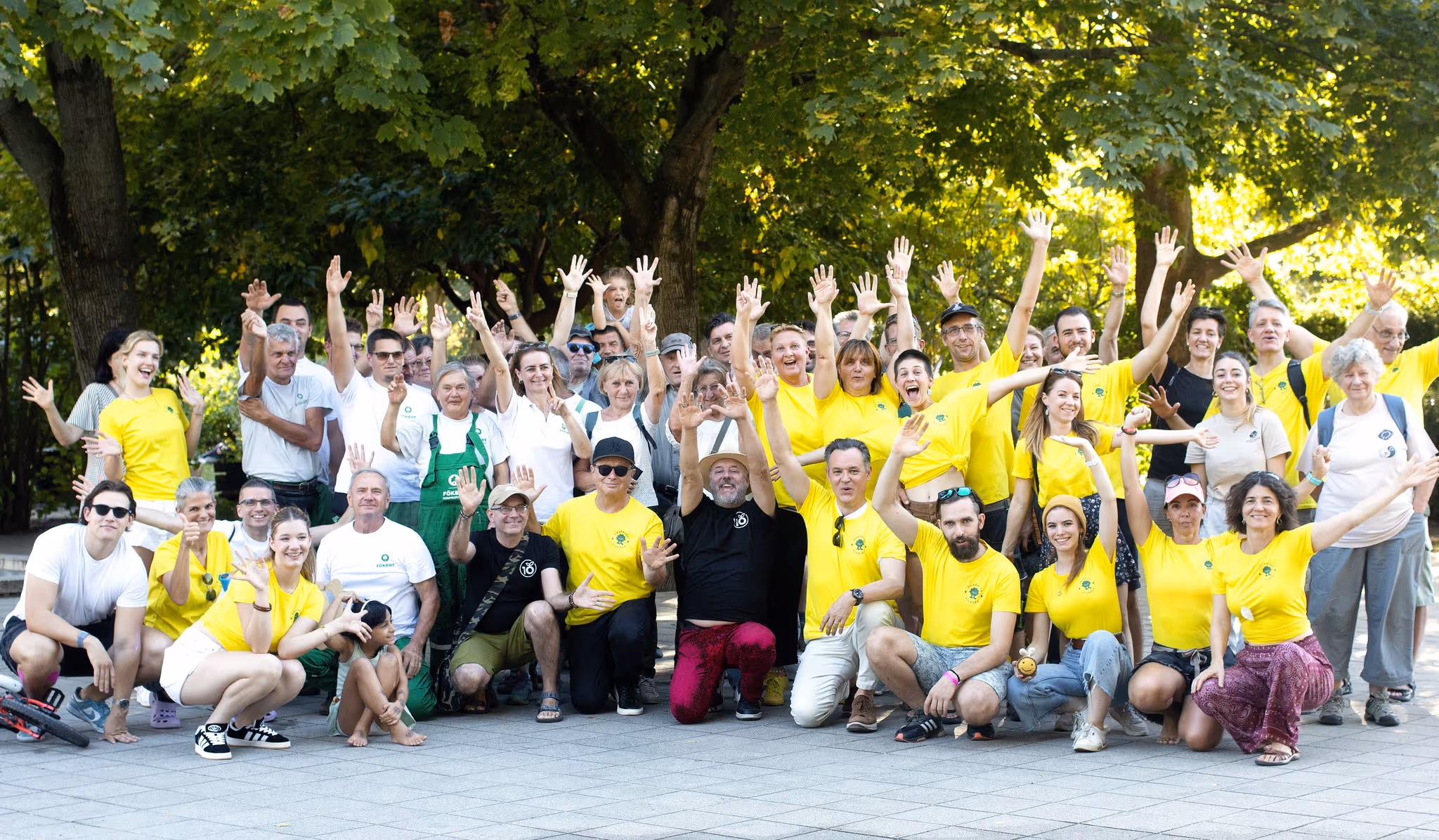 Csoportkép vidám emberekről, akik zöld fák alatt állnak, többségük sárga pólóban, kezeiket vidáman felemelve.
