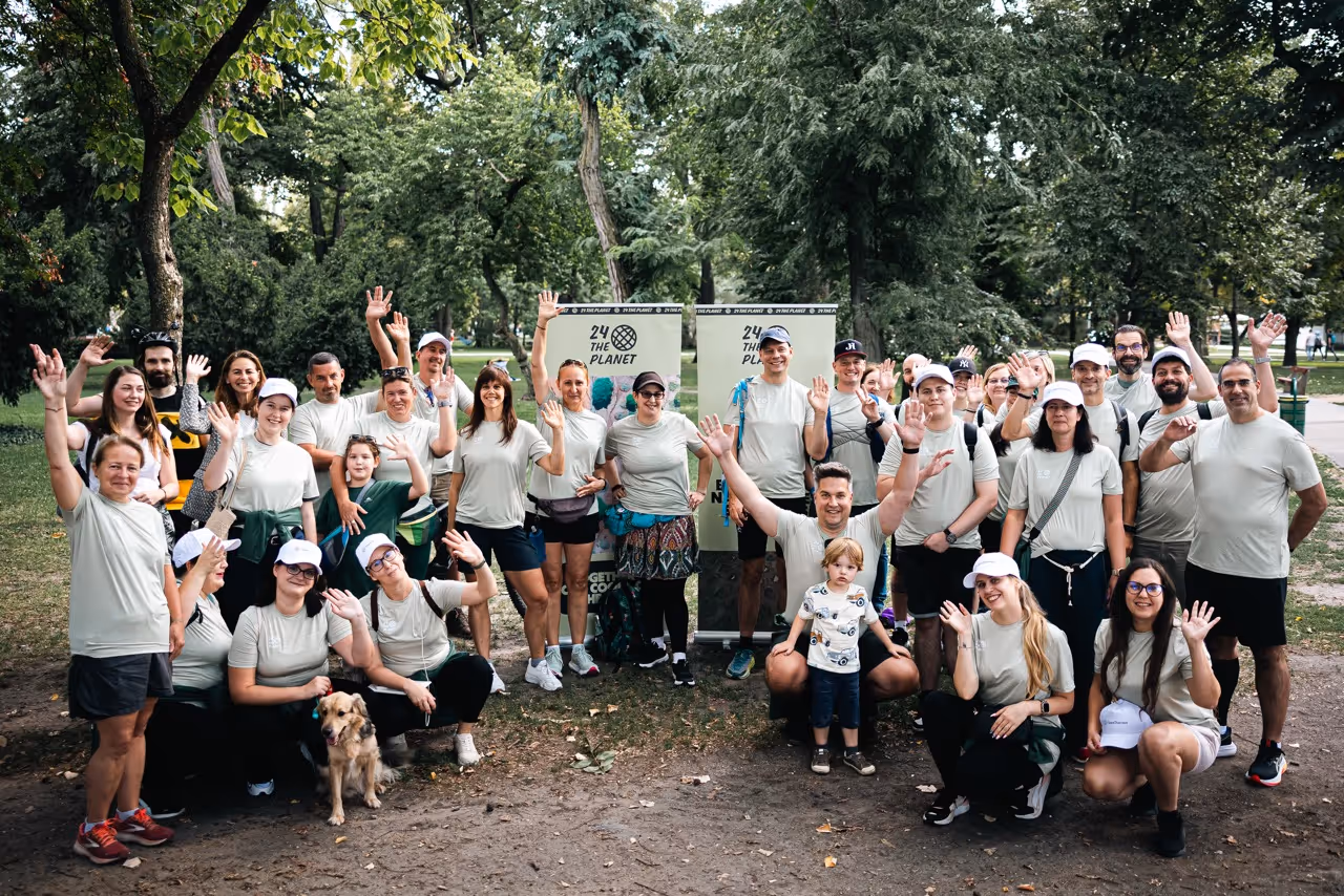 Csoport ember egy parkban, mindannyian fehér pólót viselnek, és integetnek a kamerának, mellette egy kutya is ül.