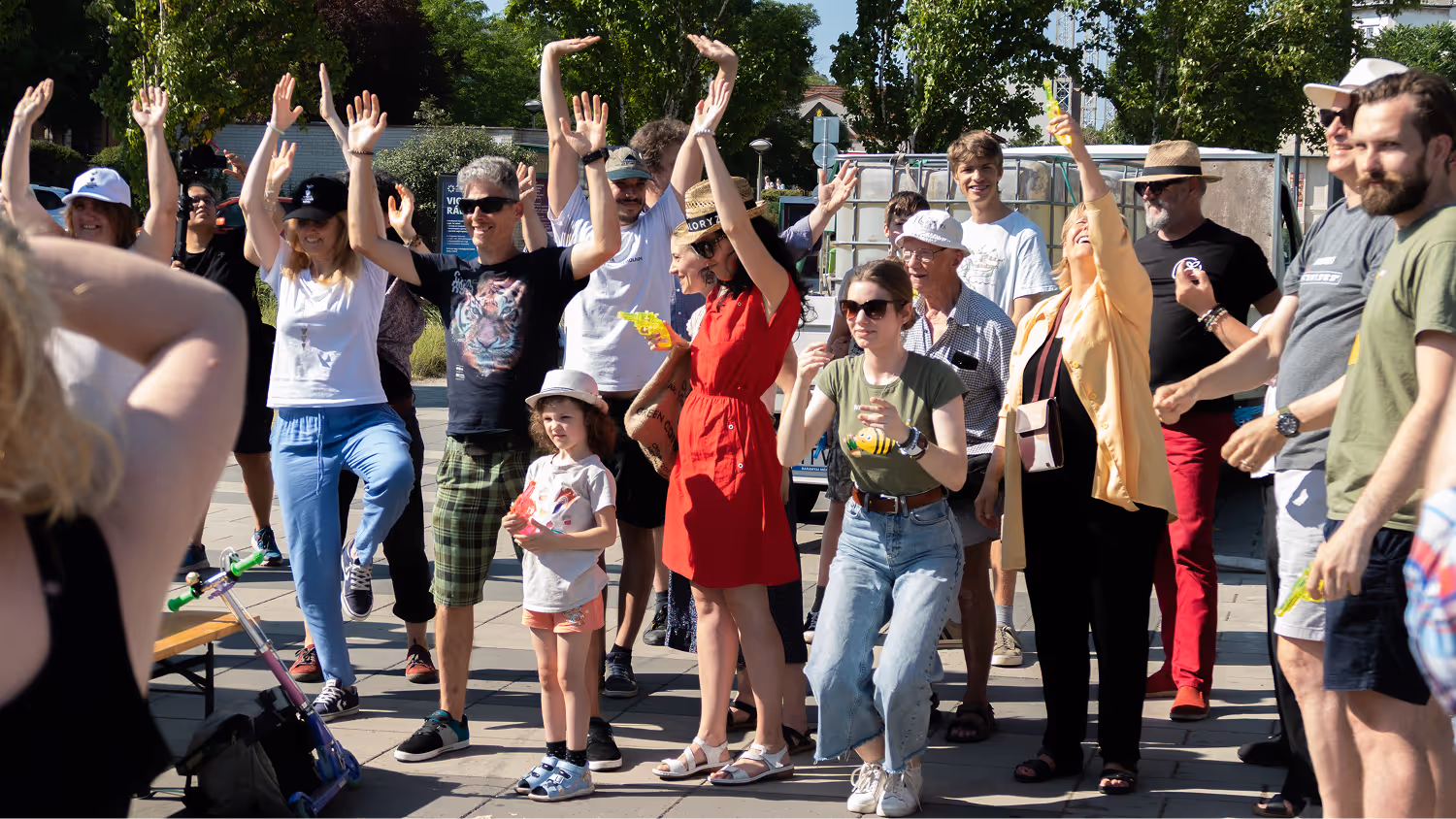 Egy csoport ember mosolyogva táncol és felkarándul egy napsütéses szabadtéri helyszínen.
