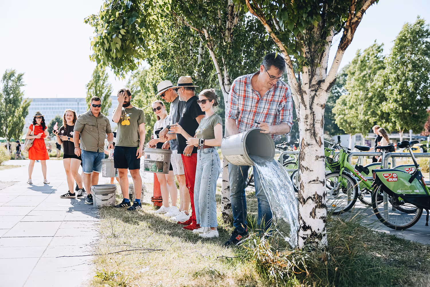 A férfi fát önt vödörből vízzel egy parkban, miközben más emberek sorban állnak mellette, napsütéses időben.