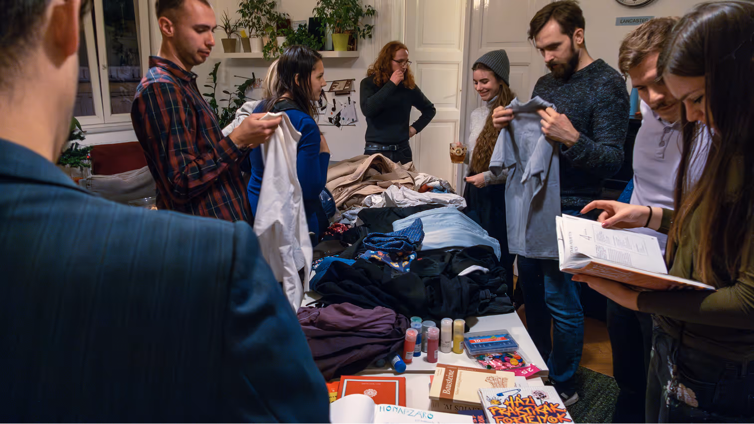 Emberek egy asztal körül ruhákat és könyveket nézegetnek egy beltéri szobában.