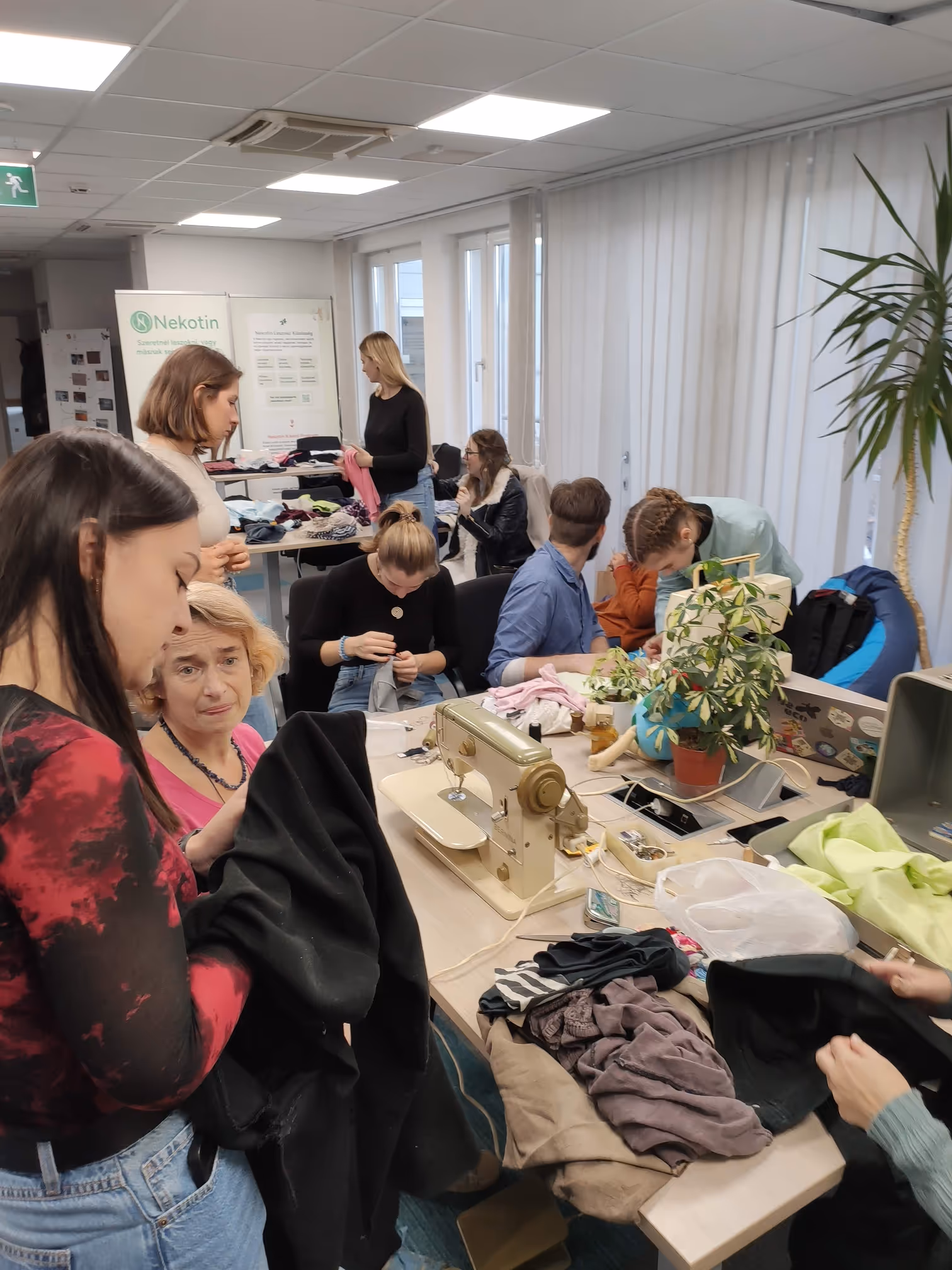 Egy csoport fiatal nő és férfi varrógép körül dolgozik és ruhákat rendez egy jól megvilágított szobában.