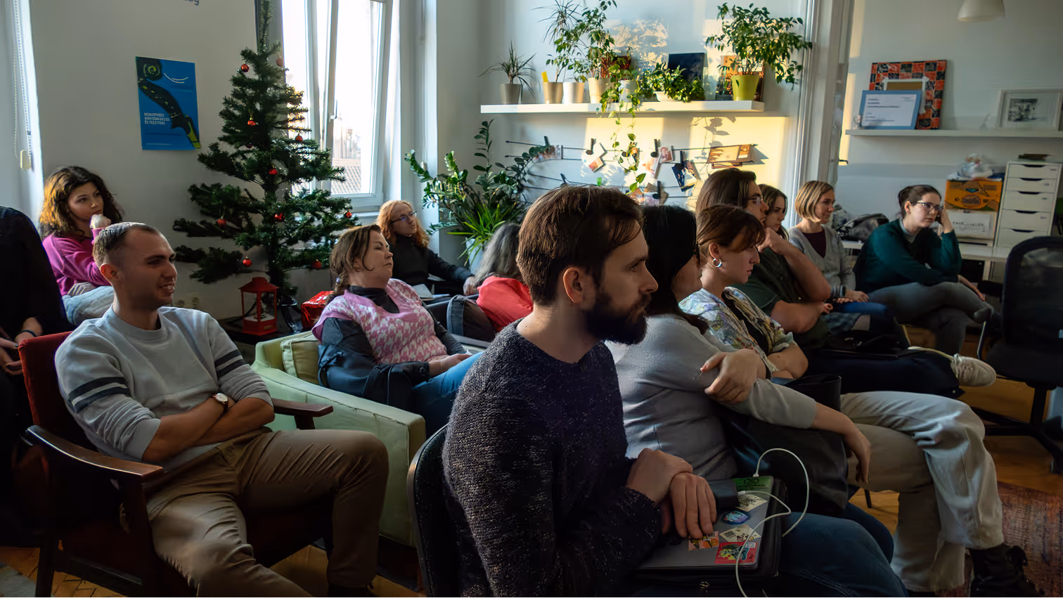 Egy csoport fiatal ember figyelmesen hallgat egy előadást egy karácsonyfával és növényekkel díszített szobában.