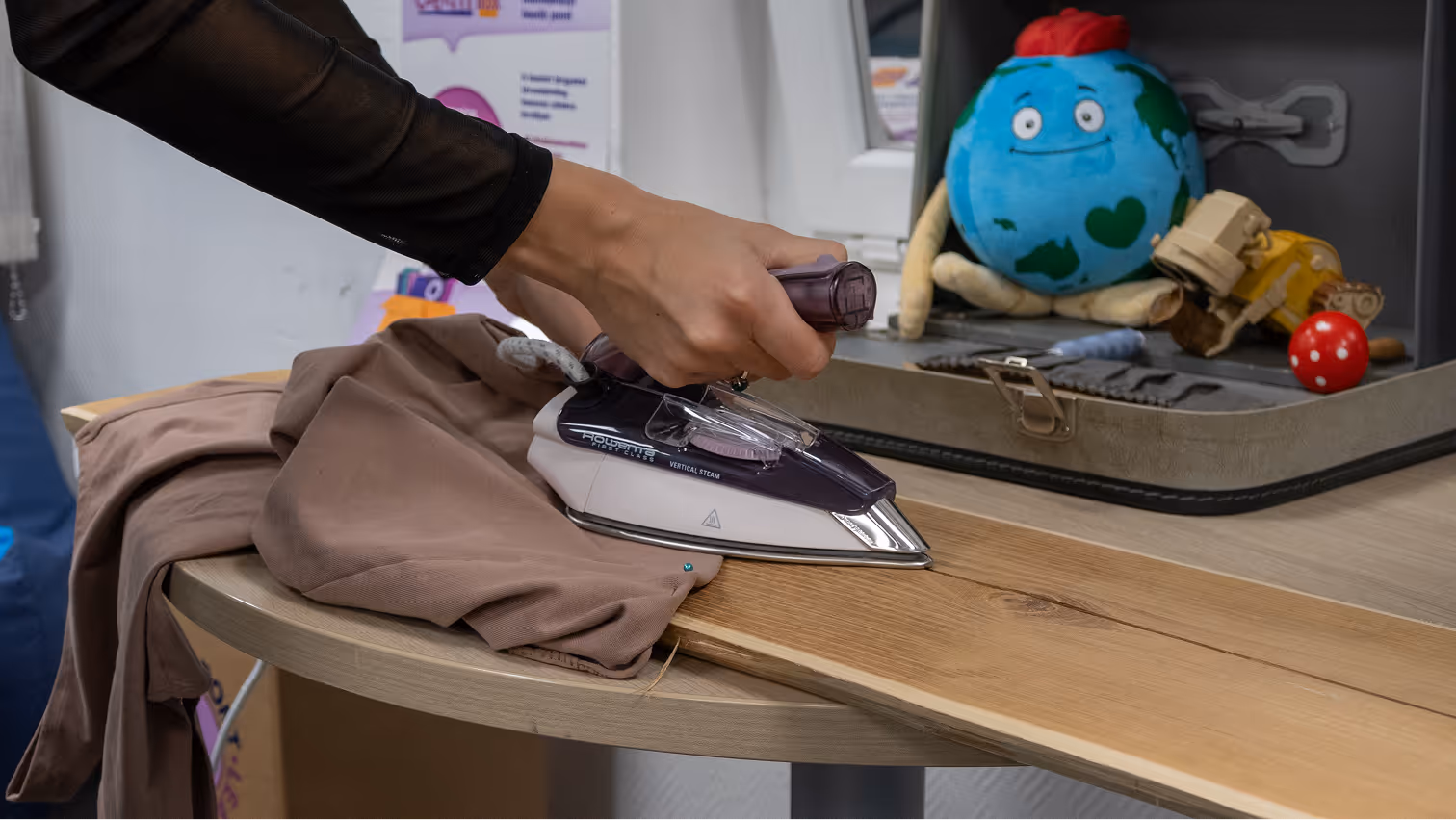 Kéz egy vasalóval barna ruhát vasal egy fa felületen egy asztalon, mellette egy nyitott bőröndben gyermekjátékok láthatók.