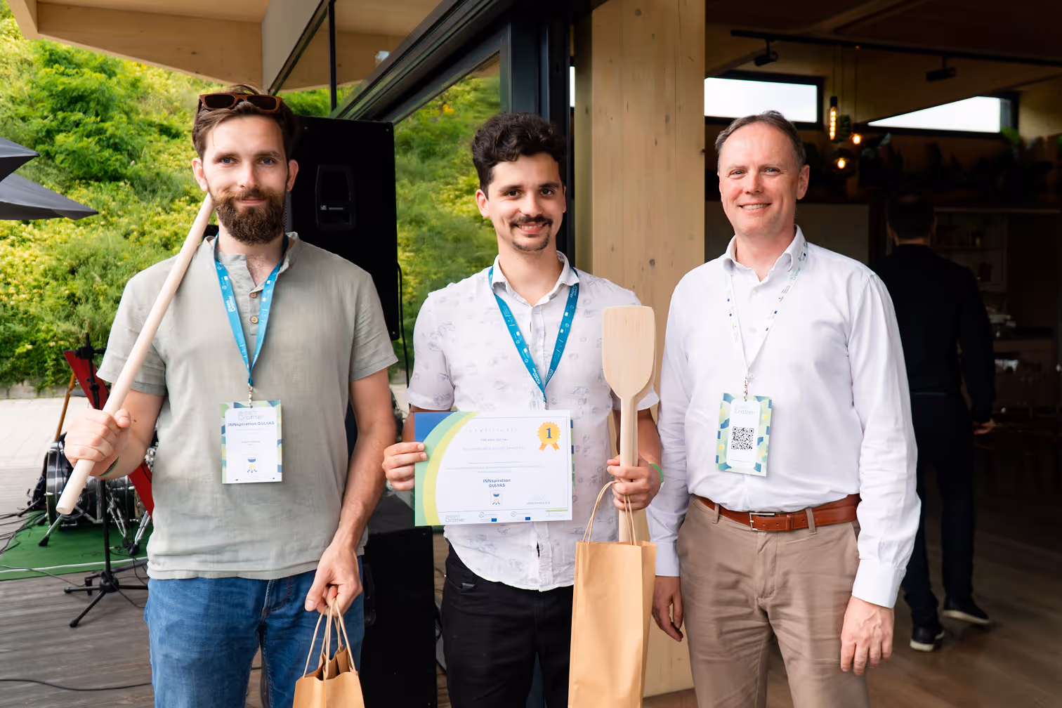 Három mosolygó férfi áll egy rendezvényen, egyikük tanúsítványt és fakanalat tart, a másik két férfi papírzacskókat és egy fakanalat.