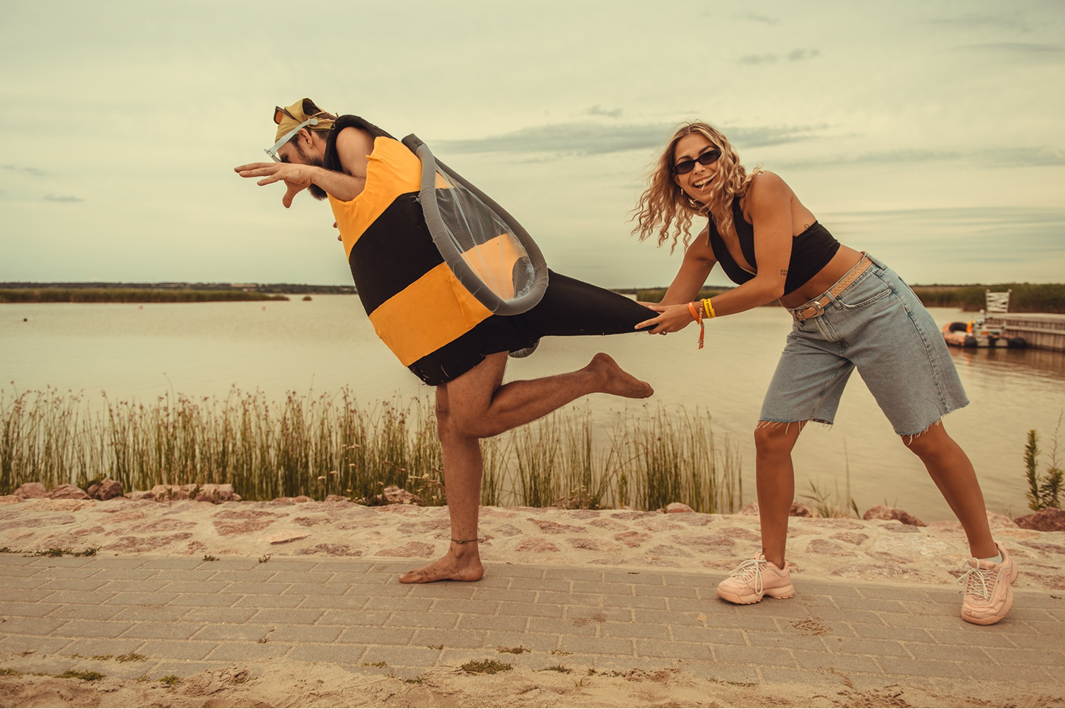 Egy nő vidáman húz egy férfit, aki méhkabátnak öltözve egy lábon áll a vízparti sétányon.