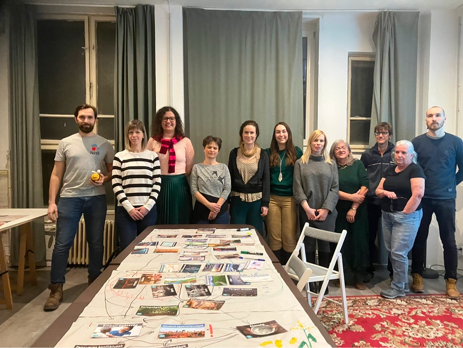 Tizenegy ember áll egy asztal mögött egy szobában, melyen különféle képek és jegyzetek vannak elrendezve.