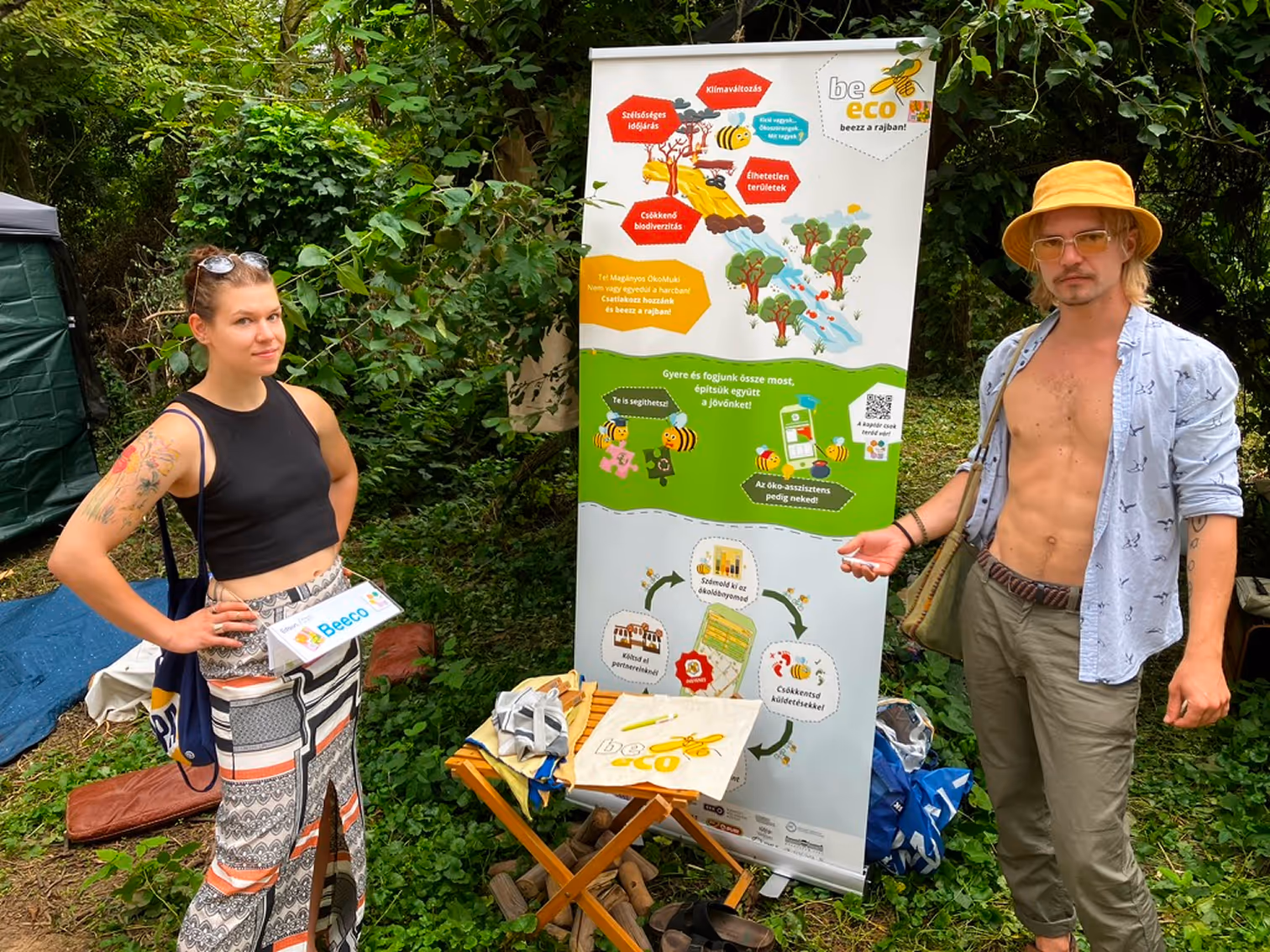 Két fiatal környezetvédelmi standnál erdei környezetben, az egyik nő fekete felsőben és mintás szoknyában, a másik férfi napsárga kalapban, ingben és zöld nadrágban áll a banner mellett.