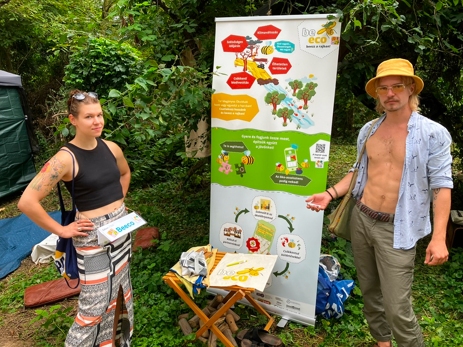 Két fiatal környezetvédelmi standnál erdei környezetben, az egyik nő fekete felsőben és mintás szoknyában, a másik férfi napsárga kalapban, ingben és zöld nadrágban áll a banner mellett.