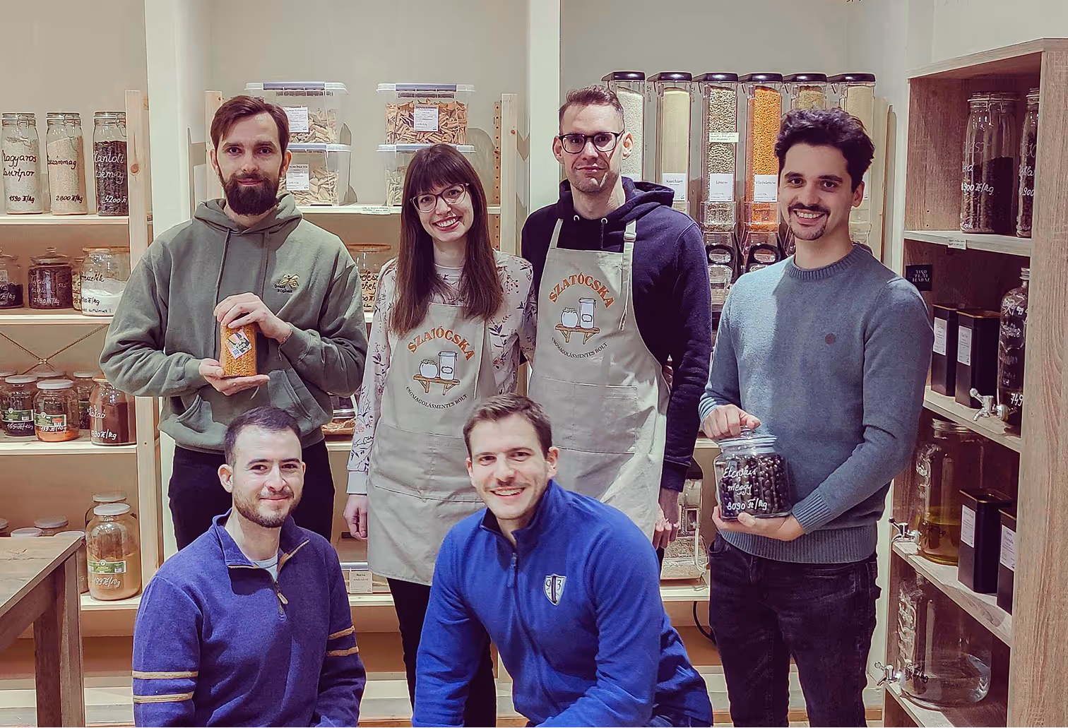 Hat mosolygó fiatal felnőtt csoportja egészséges élelmiszereket tartalmazó üveg és műanyag tárolókkal a háttérben.