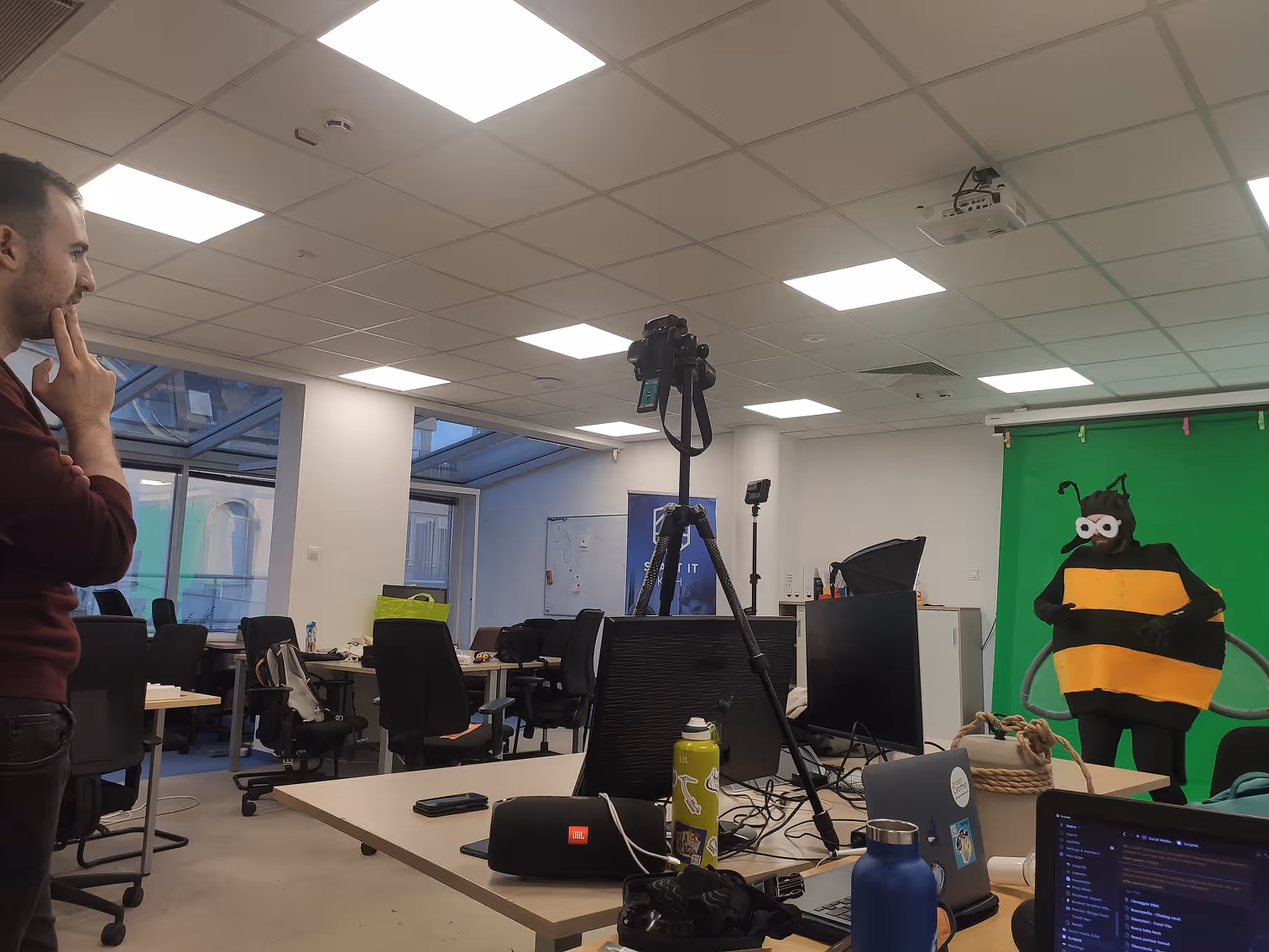 Egy férfi gondolkodik egy irodai térben, ahol valaki méhecske jelmezben áll egy zöld hátterű fotózási saroknál.