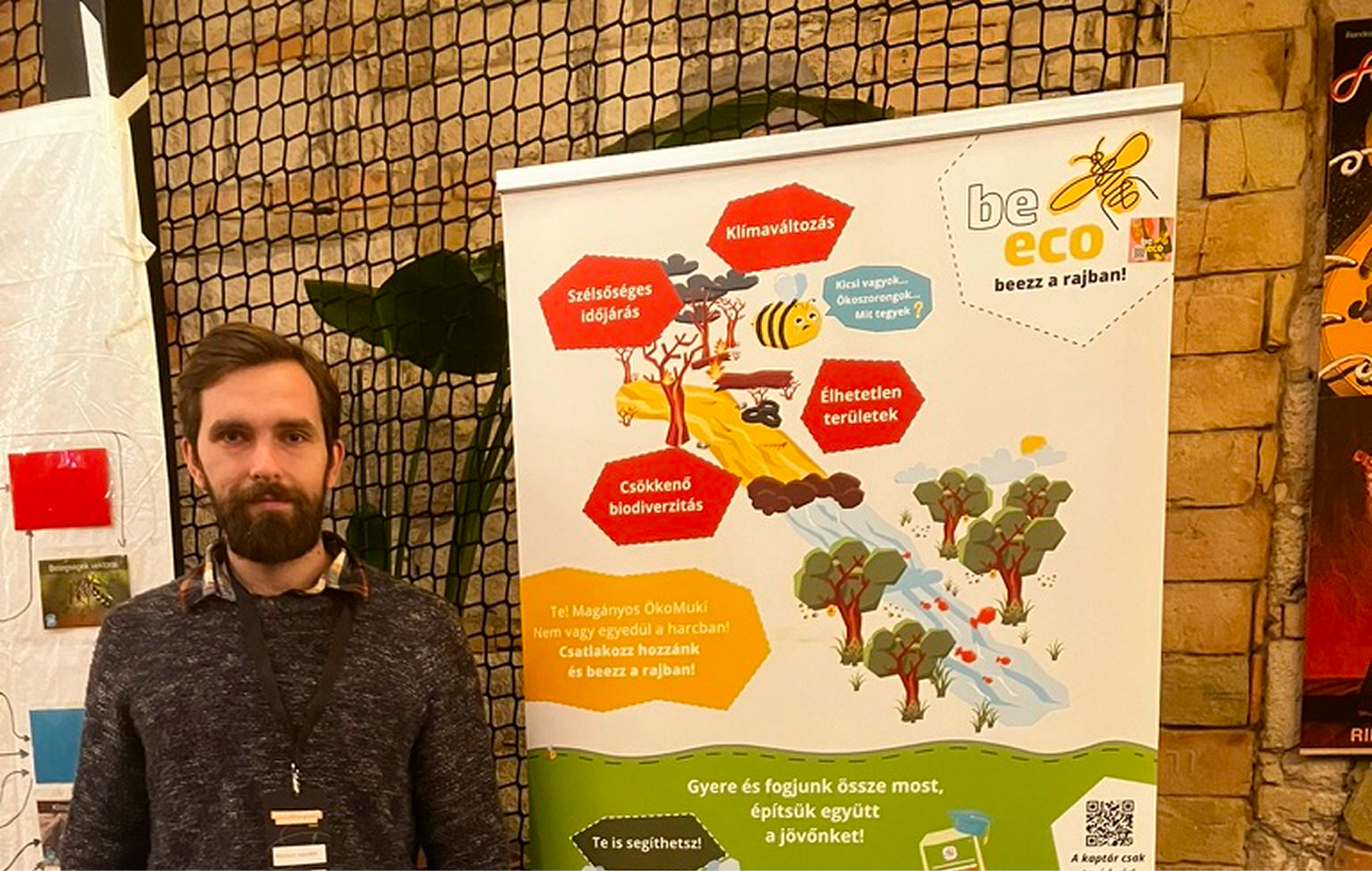 Egy férfi pózol egy hálós kerítés előtt egy környezetvédelmi témájú be eco kampány plakát mellett, amely a klímaváltozás és a biodiverzitás csökkenése veszélyeire figyelmeztet.