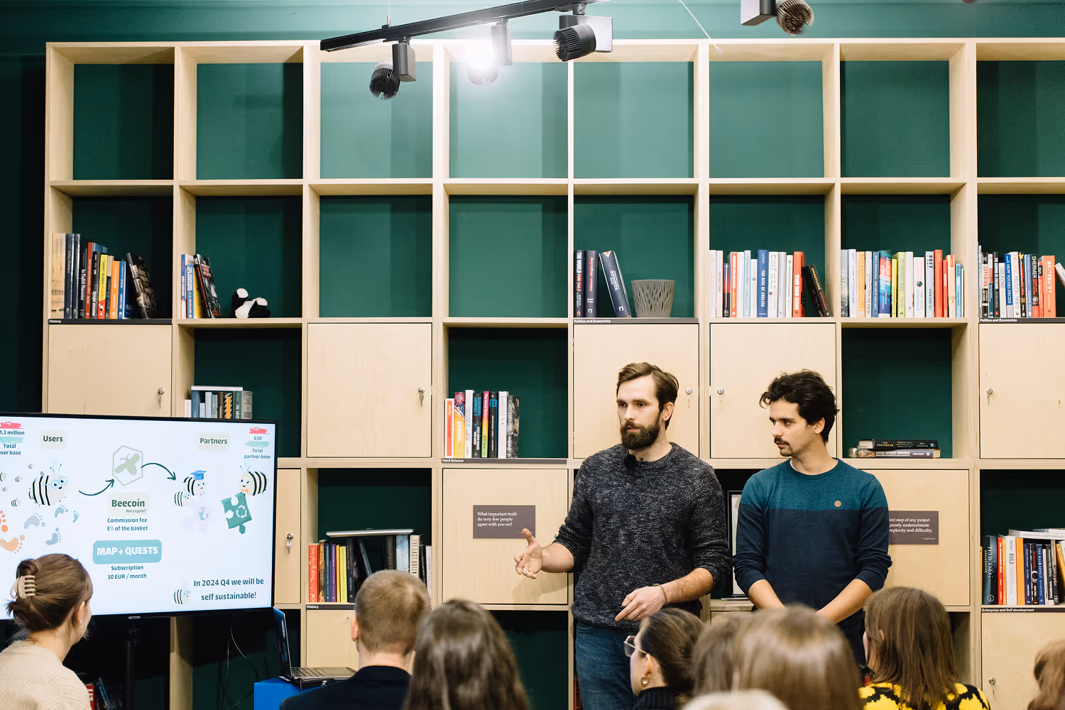 Két férfi prezentál egy előadást egy könyvespolccal teli fal előtt, miközben néhány ember hallgatja őket és egy nagy képernyőn grafikonok láthatók.