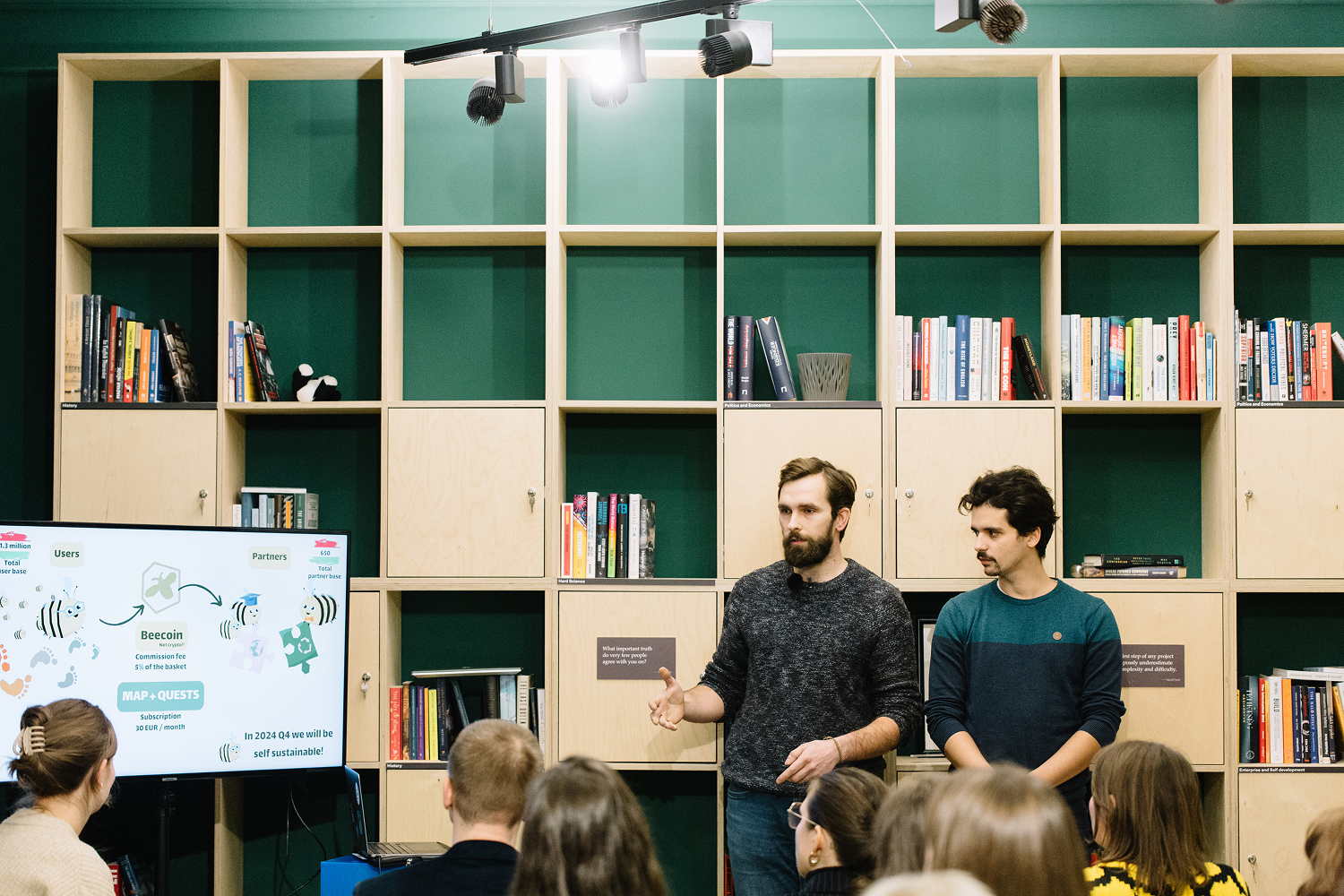 Két férfi prezentál egy előadást egy könyvespolccal teli fal előtt, miközben néhány ember hallgatja őket és egy nagy képernyőn grafikonok láthatók.