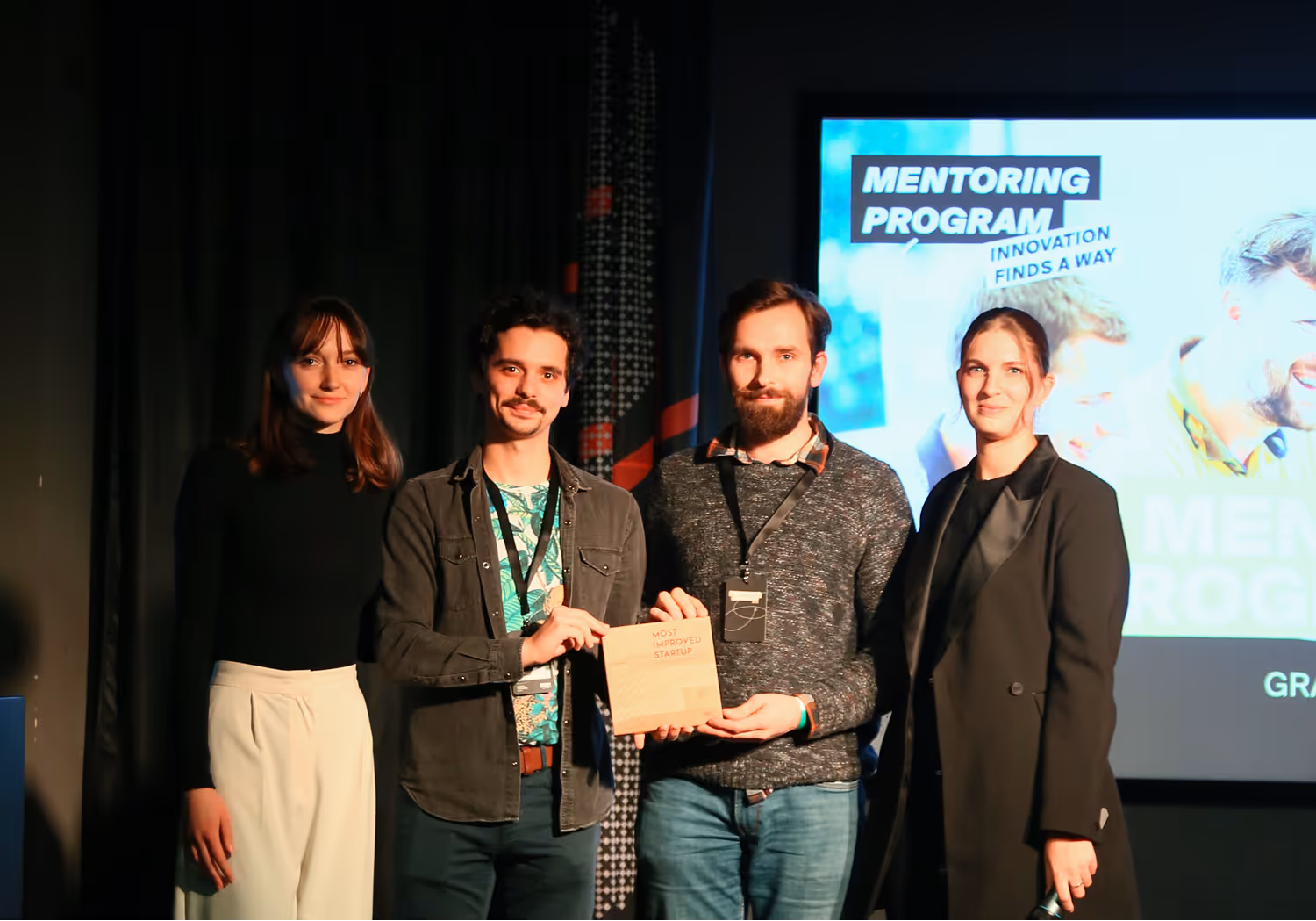 Négy fiatal ember áll színpadon, az egyikük egy fa díjat tart, amelyen az áll: "Most Improved Startup"; a háttérben "Mentoring Program" felirat látható.
