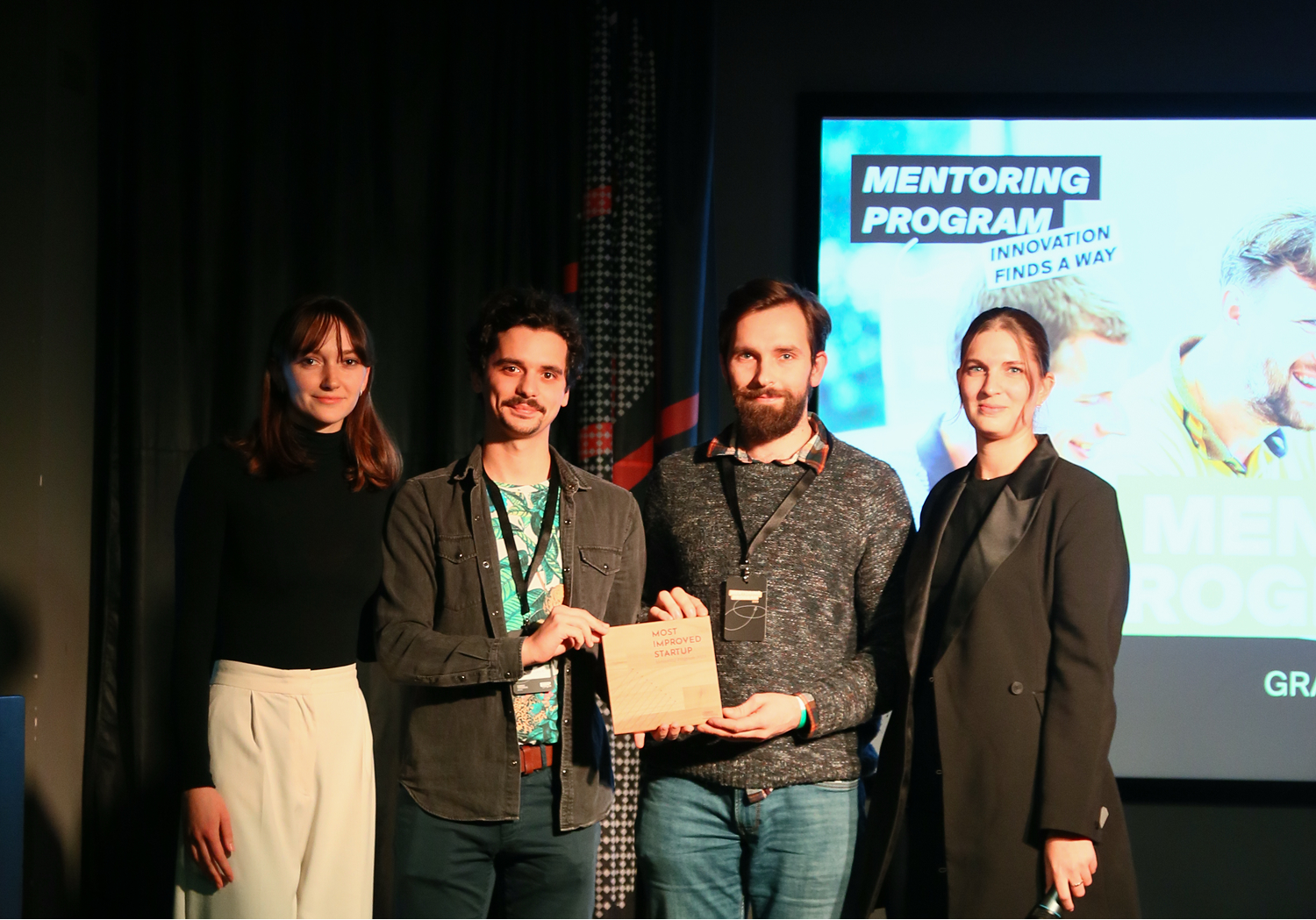 Négy fiatal ember áll színpadon, az egyikük egy fa díjat tart, amelyen az áll: "Most Improved Startup"; a háttérben "Mentoring Program" felirat látható.
