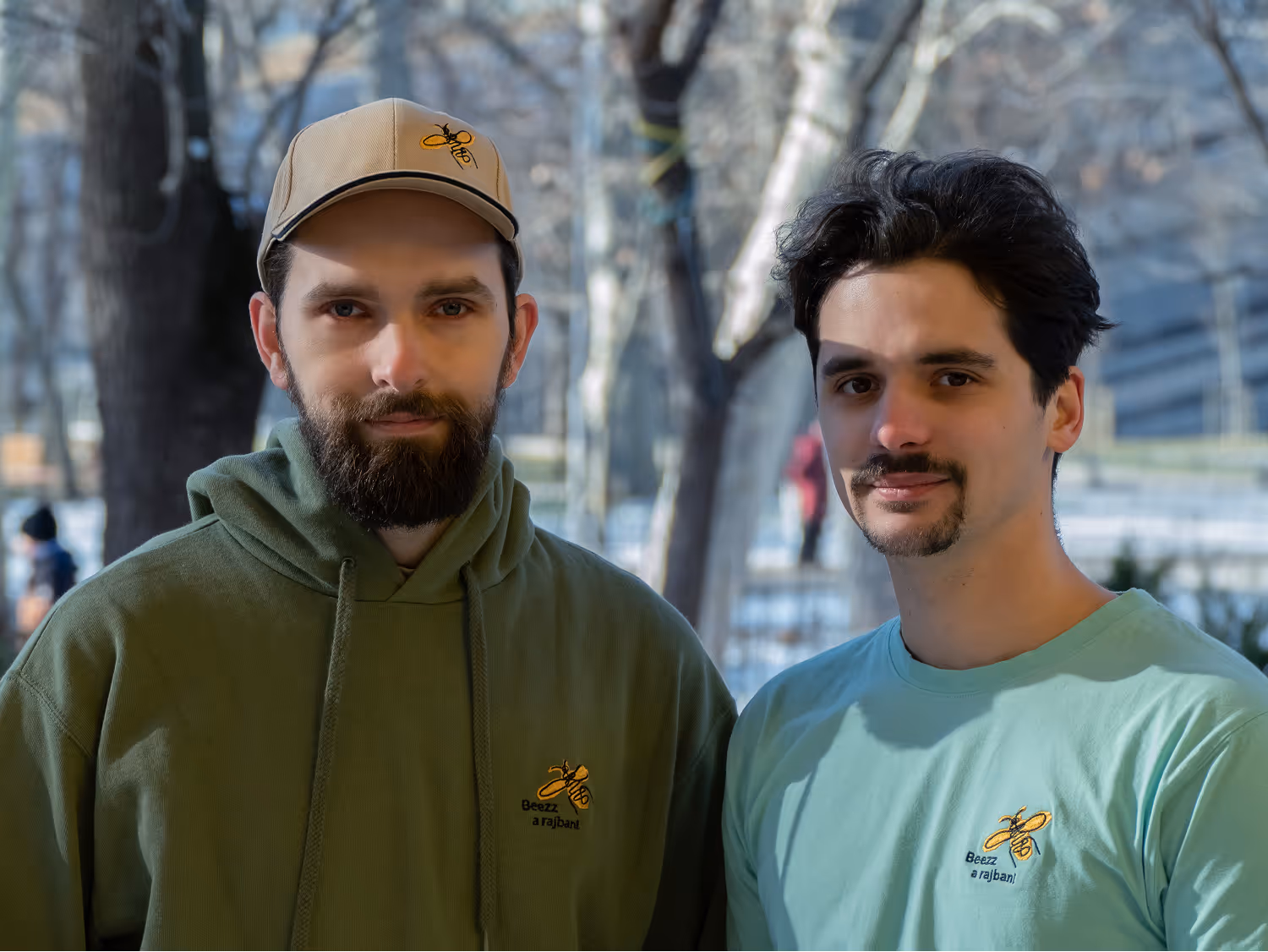 Két férfi áll egymás mellett kint a szabadban, egyiket zöld kapucnis pulóver és sapka, a másikat világoskék póló visel, mindkettőn sárga méh logóval és magyar felirattal.