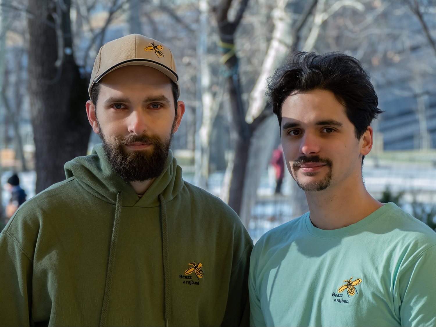 Két férfi áll egymás mellett kint a szabadban, egyiket zöld kapucnis pulóver és sapka, a másikat világoskék póló visel, mindkettőn sárga méh logóval és magyar felirattal.