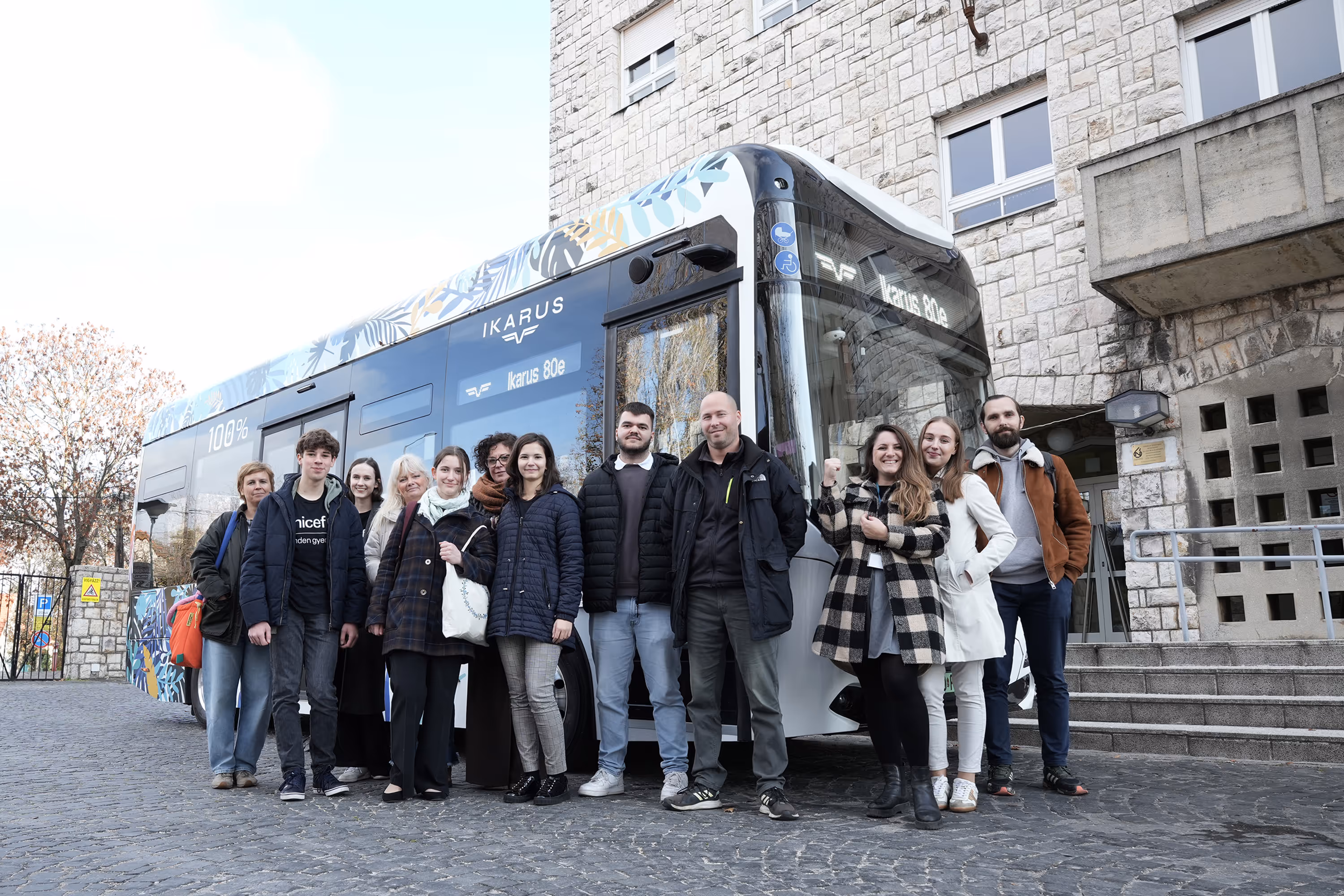Tizenkét ember áll egy Ikarus 80e elektromos busz előtt egy kőfalú épület mellett.
