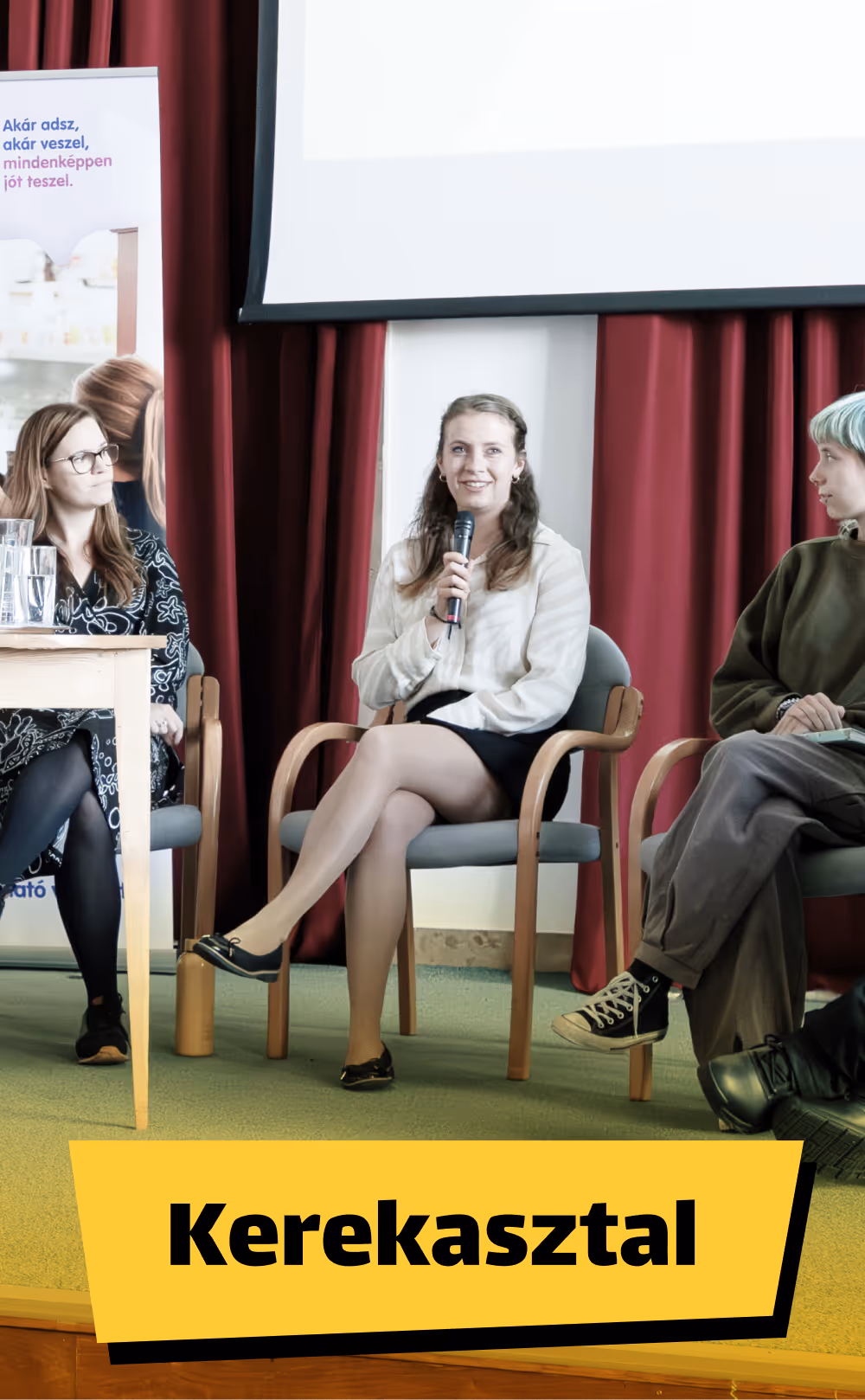 Három nő ül egy kerekasztal-beszélgetésen, az egyik mikrofont tartva beszél.