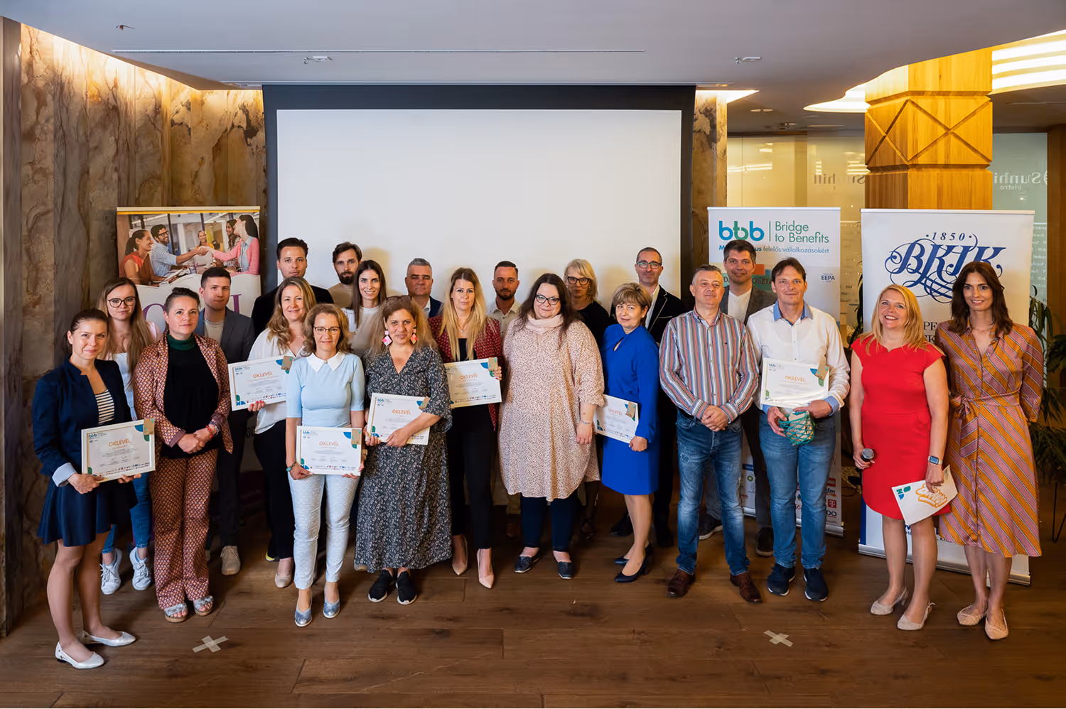 Csoportkép 22 felnőtt résztvevőről, akik egy rendezvényen okleveleket tartanak egy konferencia terem előtt.