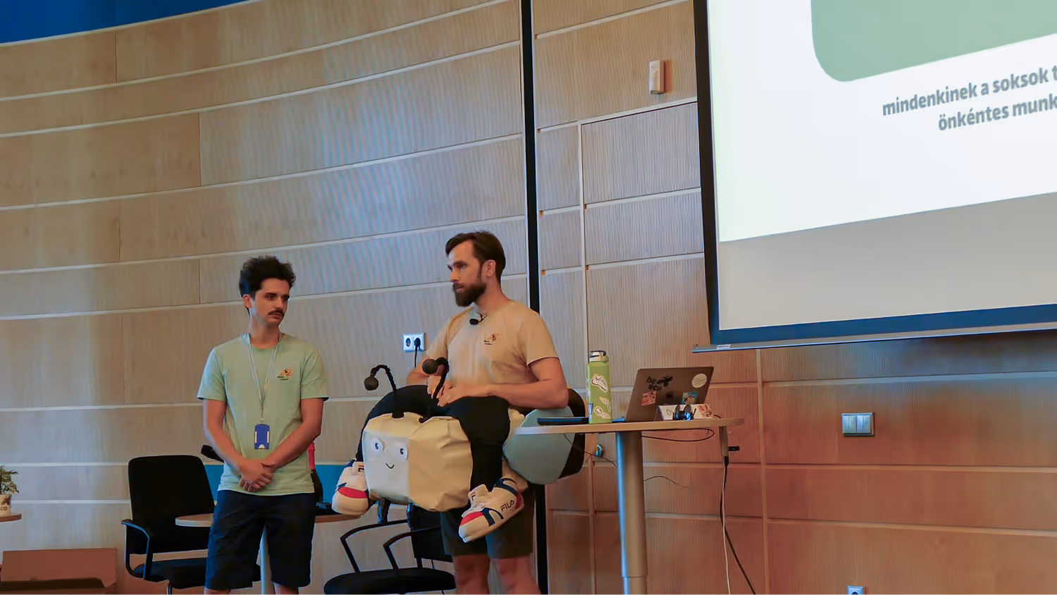 Két férfi áll egy előadóteremben, az egyik egy méh jelmezben, a másik zöld pólót és nyakba akasztható kártyát visel.