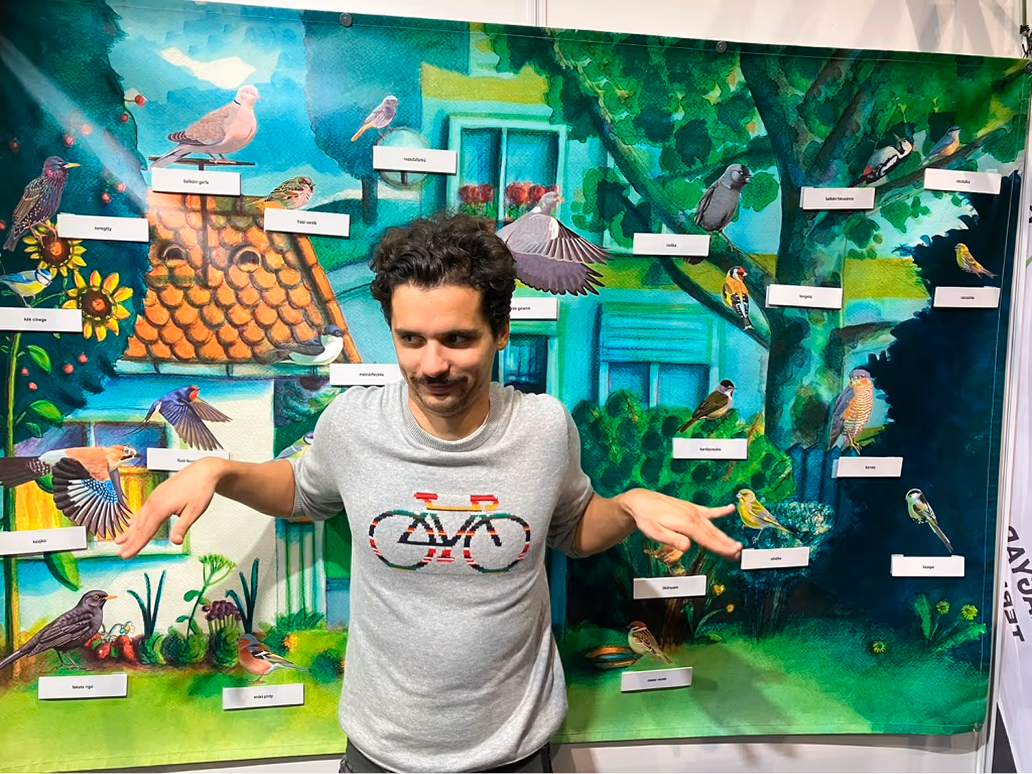 Fiatal férfi szürke pulóverben, bicikli mintával, madárillusztrációk előtt áll, melyek nevei magyarul vannak feltüntetve.