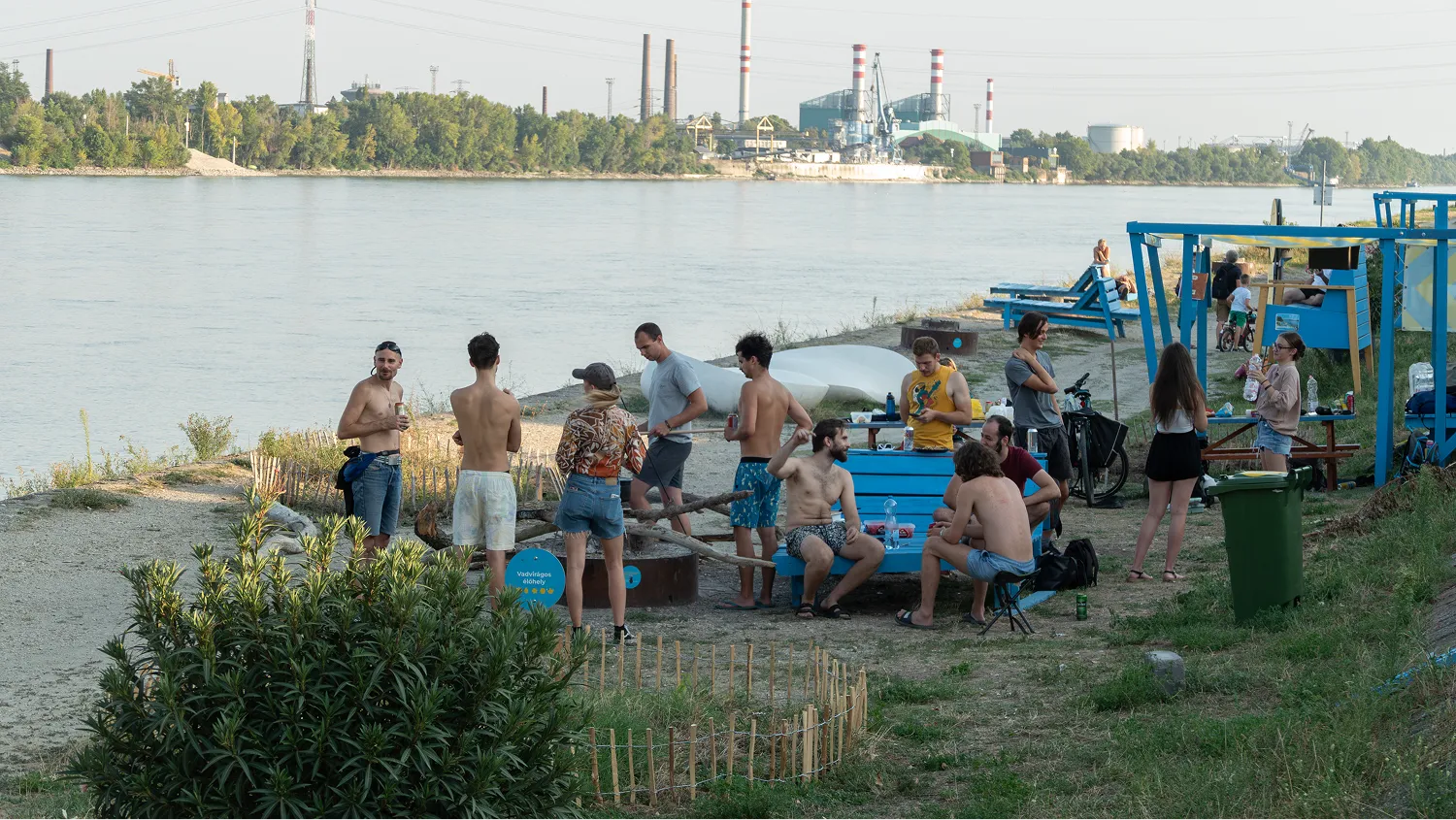Emberek csoportja beszélget és pihen a folyóparti szabadban, háttérben ipari épületekkel.