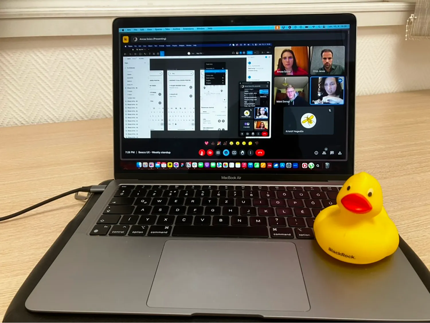 MacBook Air laptop on a wooden desk displaying a virtual meeting with multiple participants and a yellow rubber duck with BlackRock logo on the keyboard.