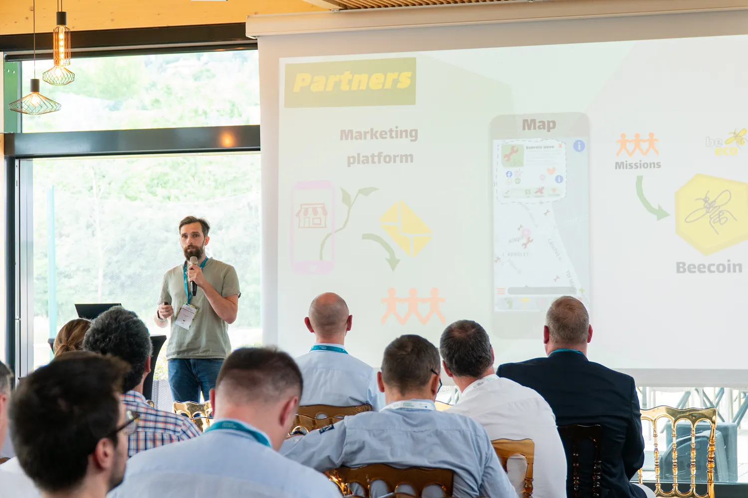 Férfi prezentációt tart mikrofonnal egy fehér vászon előtt, ahol egy marketing platform és Beecoin térkép diagram látható, a közönség arccal felé néz.