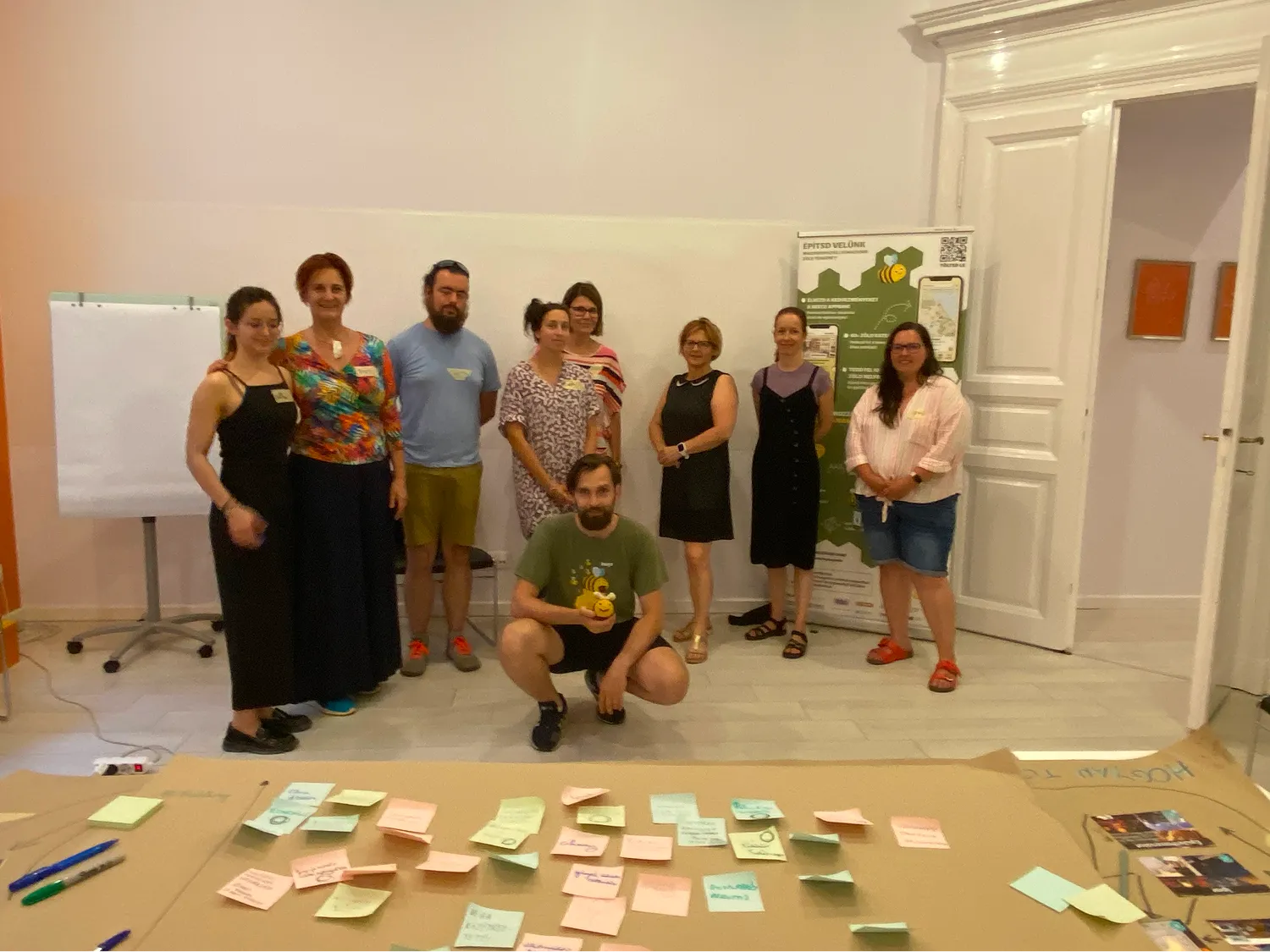 Nyolc ember egy konferencia teremben áll és egy férfi guggol előttük, a padlón több színes post-it cetli szétszórva.