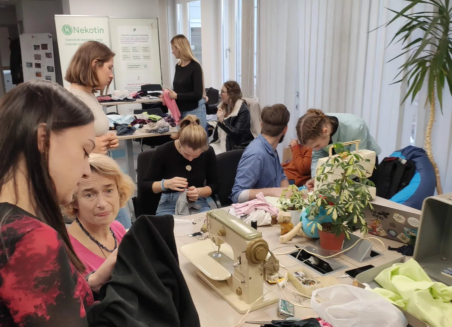 Csoport emberek egy szabóműhelyben, varrógép, ruhák és növények a asztalon, ahol egy nő varr, míg mások beszélgetnek és nézelődnek.
