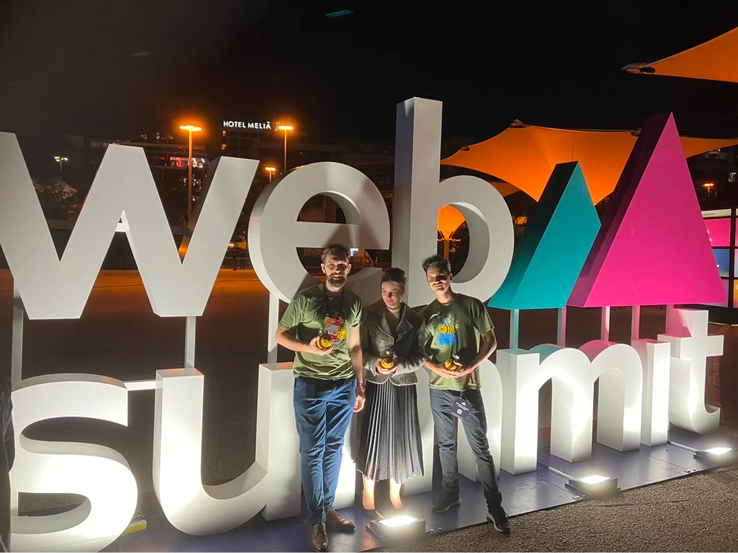 Három fiatal áll egy világító Web Summit felirat előtt éjszaka, mindhárman kezükben apró tárgyat tartanak, mögöttük hotel és narancssárga napellenzők láthatók.