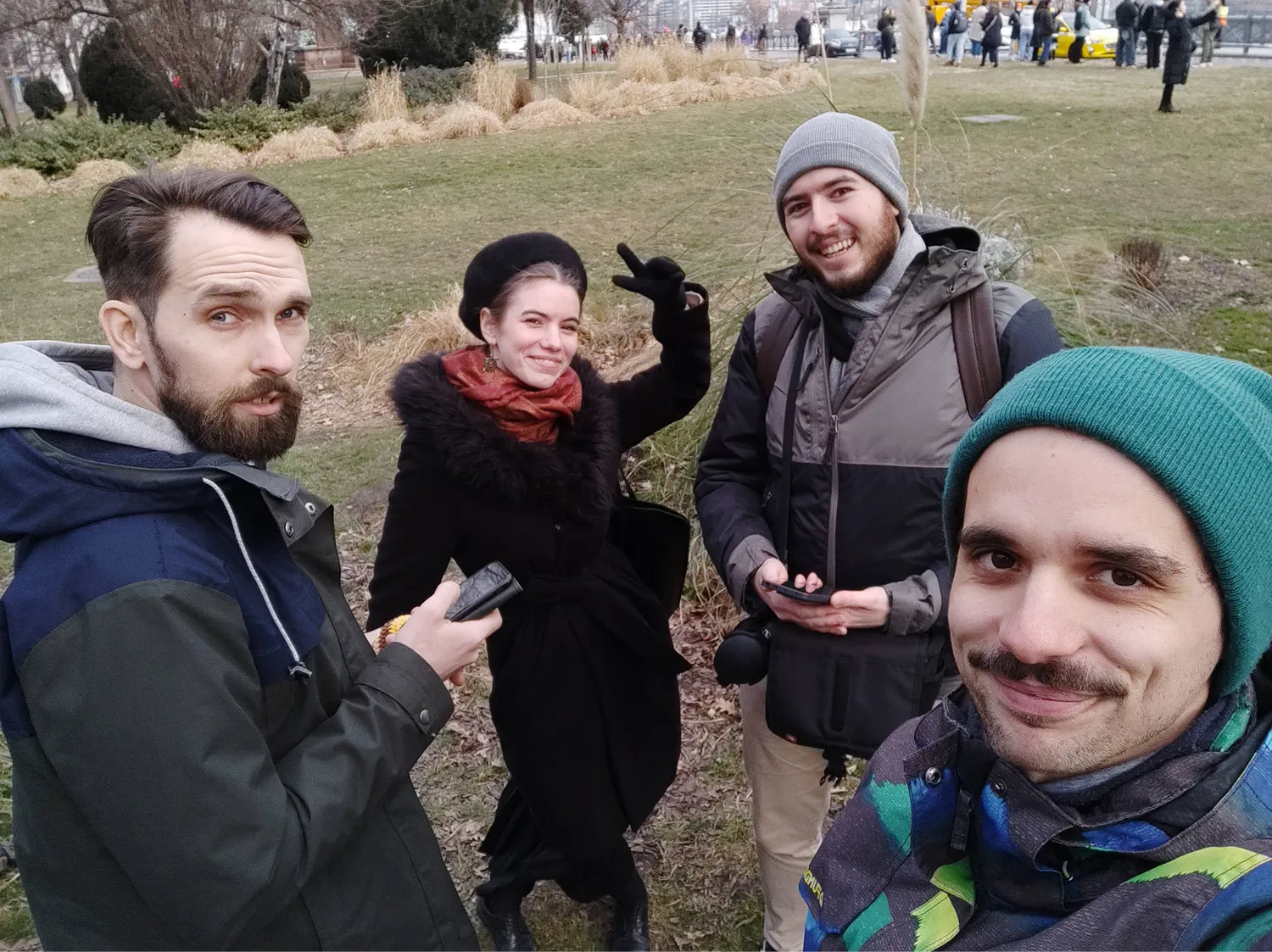 Négy fiatal áll egy parkban téli ruhában, közülük az egyik békés jelzést mutat és mosolyog.