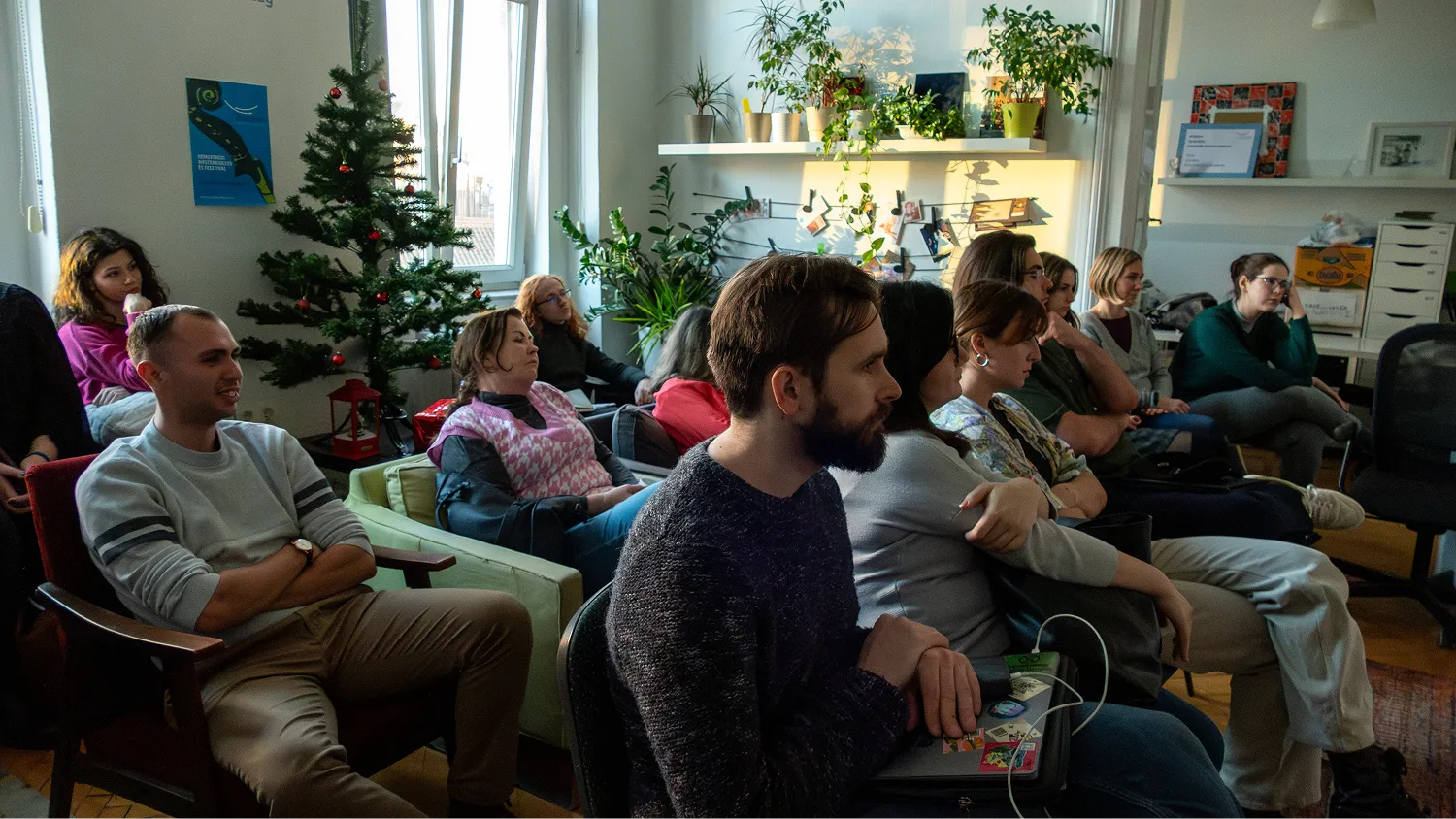 Csoport fiatal felnőtt hallgató ül egy nappaliban, figyelmesen hallgatnak egy bemutatót vagy beszélgetést, karácsonyfa és növények díszítik a teret.