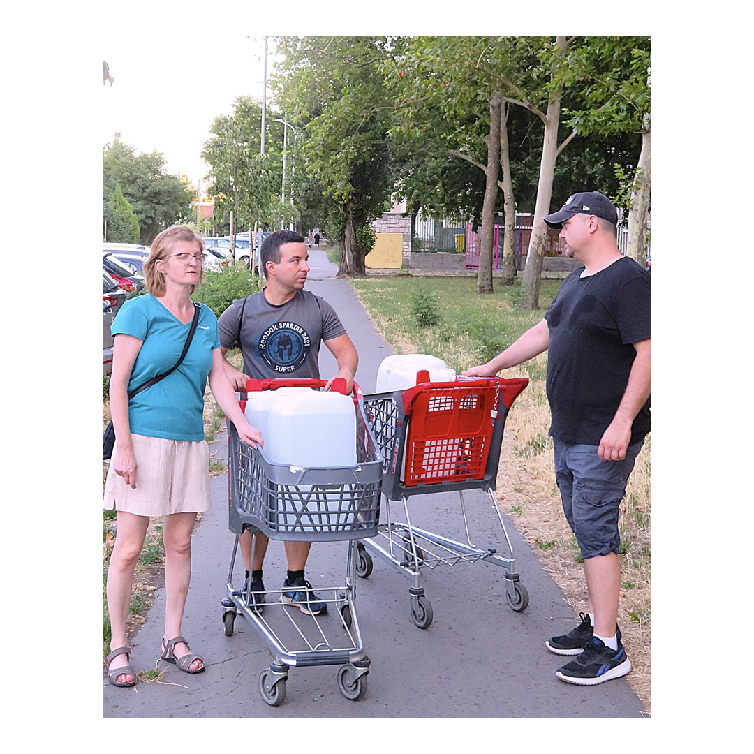 Három ember bevásárlókocsikkal az utcán, amikben nagy fehér kannák vannak.