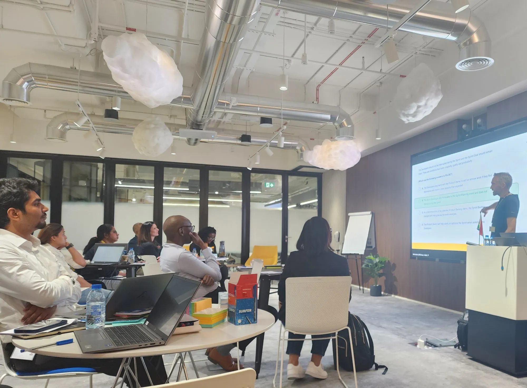 Agile coach leading a live training session, presenting to a seated group in a bright, plant-filled workshop space